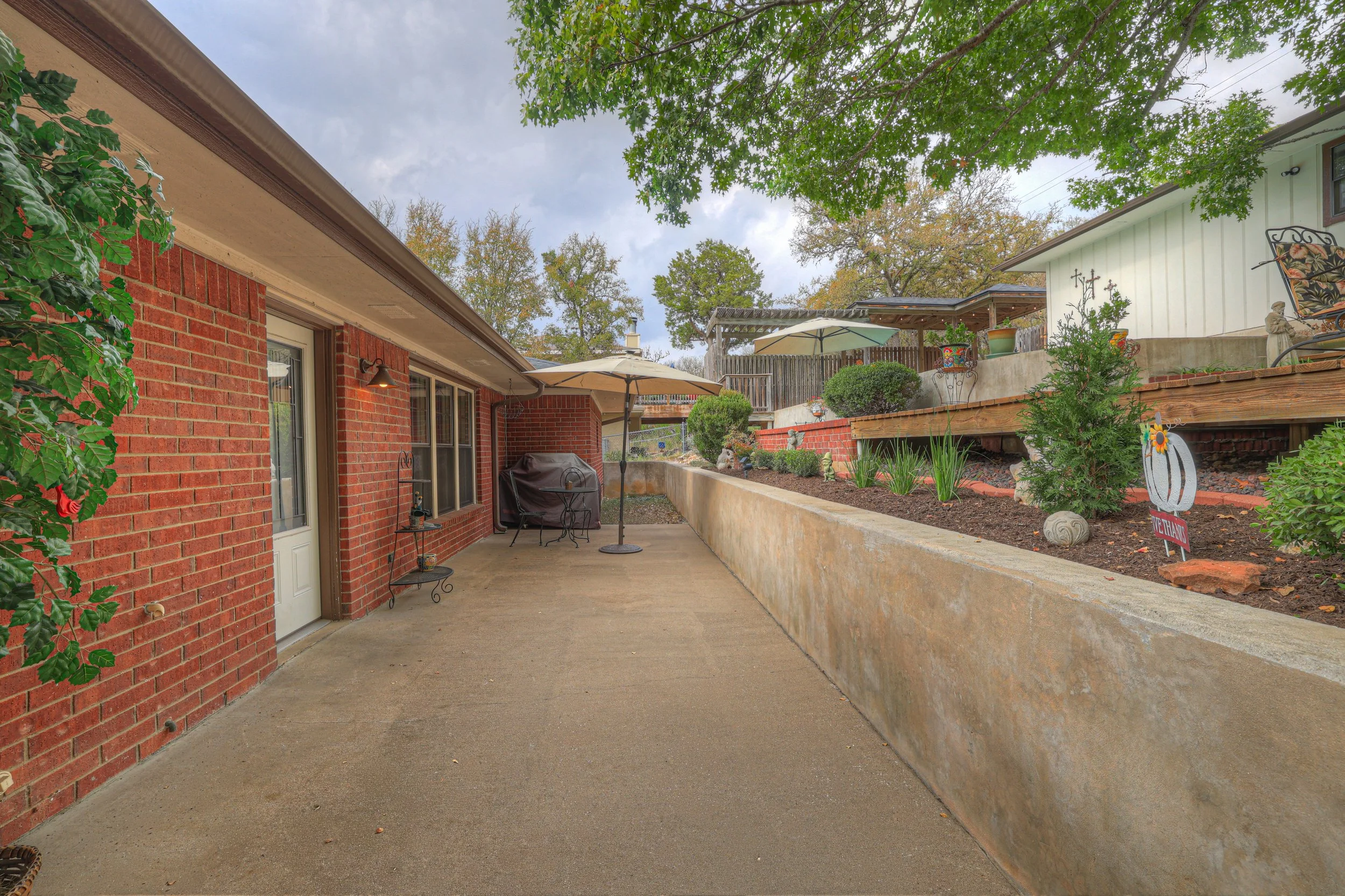 Backyard patio with concrete floor, brick house wall, patio furniture including umbrella, barbecue grill, and potted plants, with a raised garden bed and trees in the background.