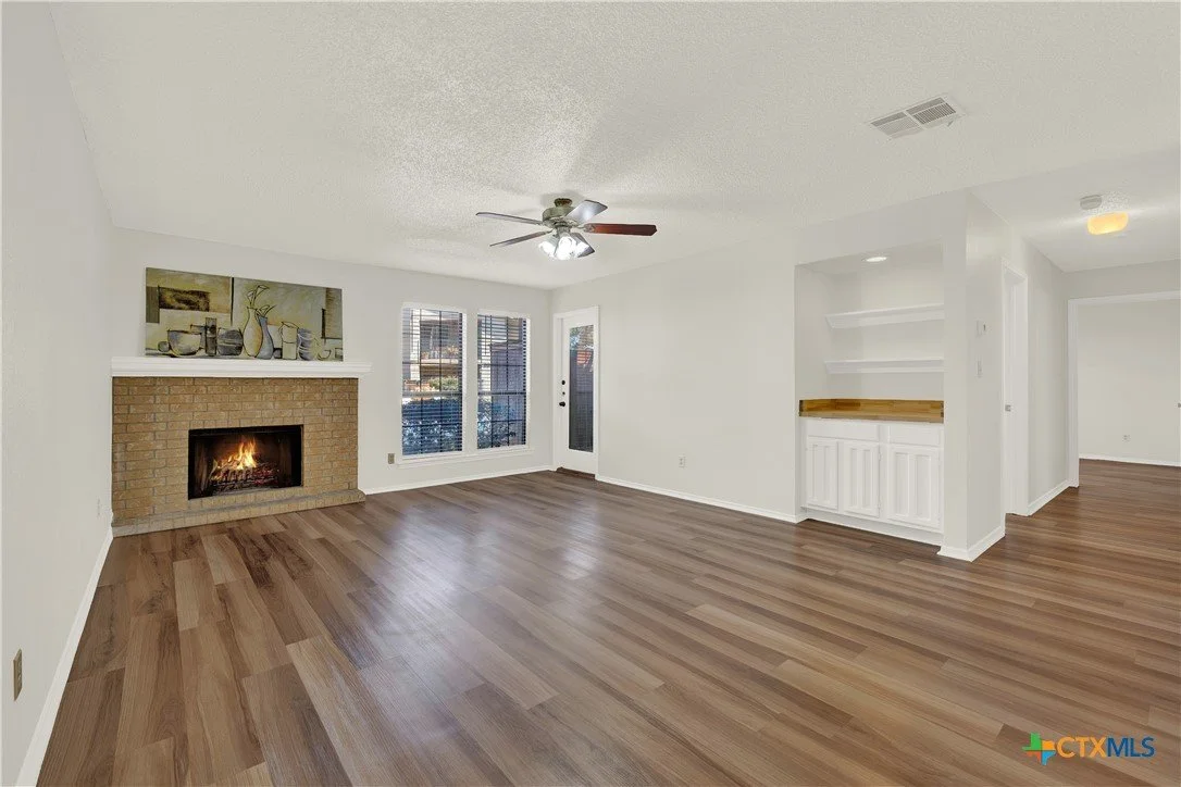 Empty living room with wood floor, white walls, a brick fireplace, a ceiling fan, and windows with blinds.