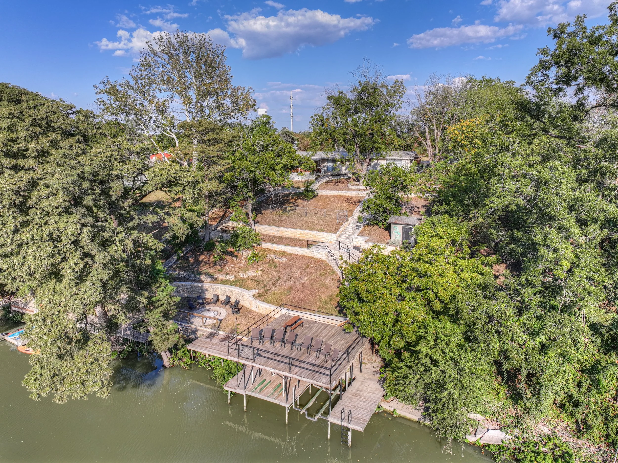 Aerial view of a lakeside area with trees, a wooden deck with chairs, and stairs leading up a hill with more trees and buildings, under a partly cloudy sky.