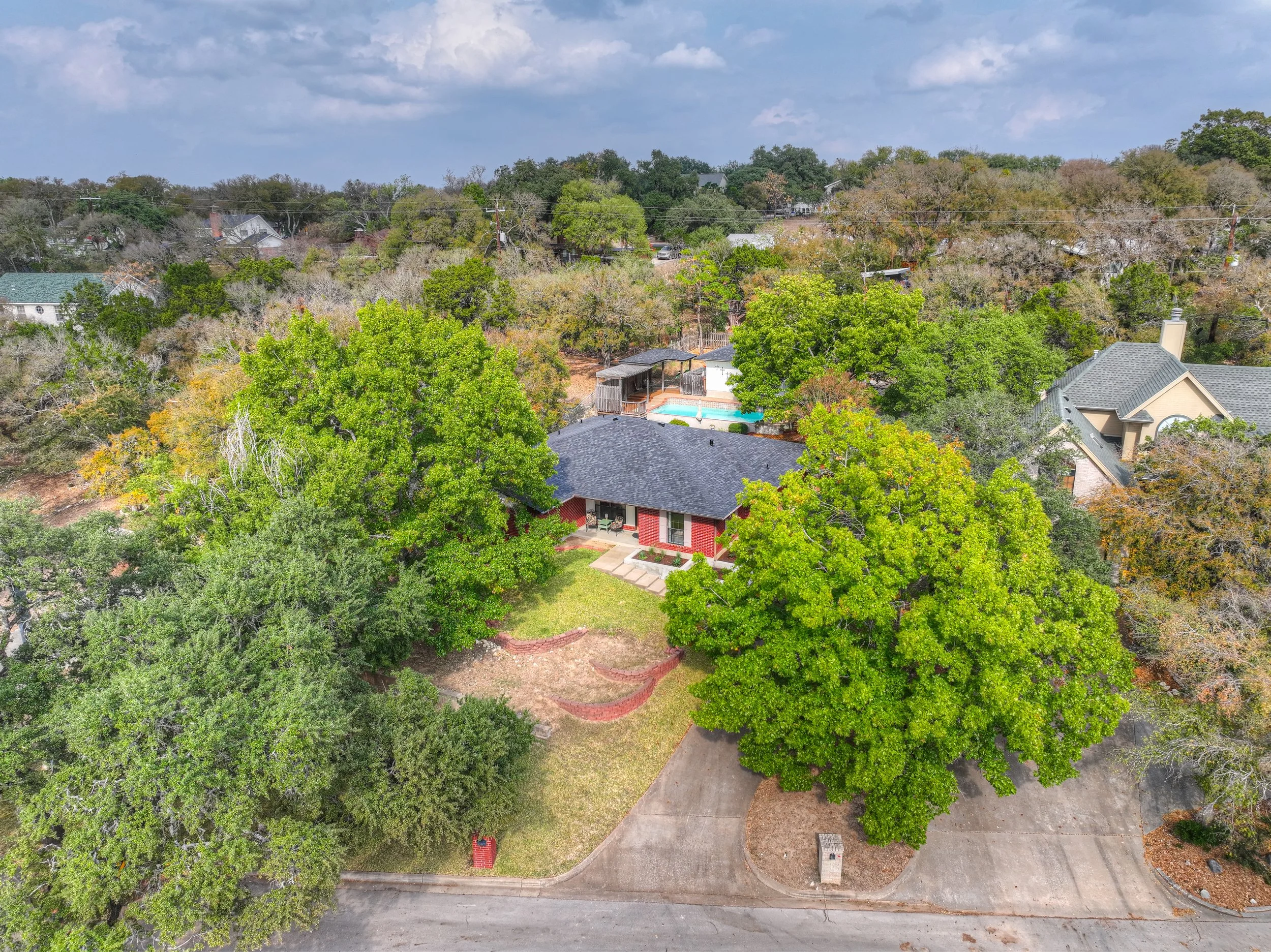 Aerial view of a residential house surrounded by large green trees, with a neighboring house visible nearby and a backyard with a small pool and a shed.