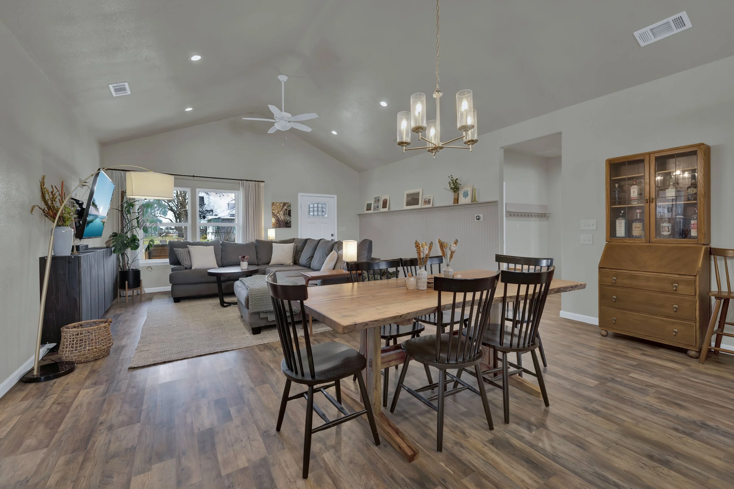 Open-concept living and dining room with a gray sectional sofa, wooden dining table with six black chairs, a wooden cabinet, and various decorative items, lit by a chandelier and natural light from windows.