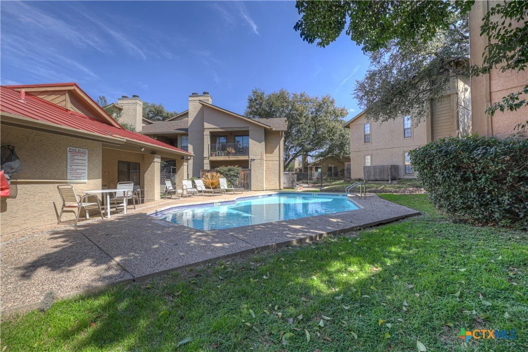 Apartment complex outdoor swimming pool with chairs and tables, surrounded by buildings, trees, and a grassy area under a clear blue sky.