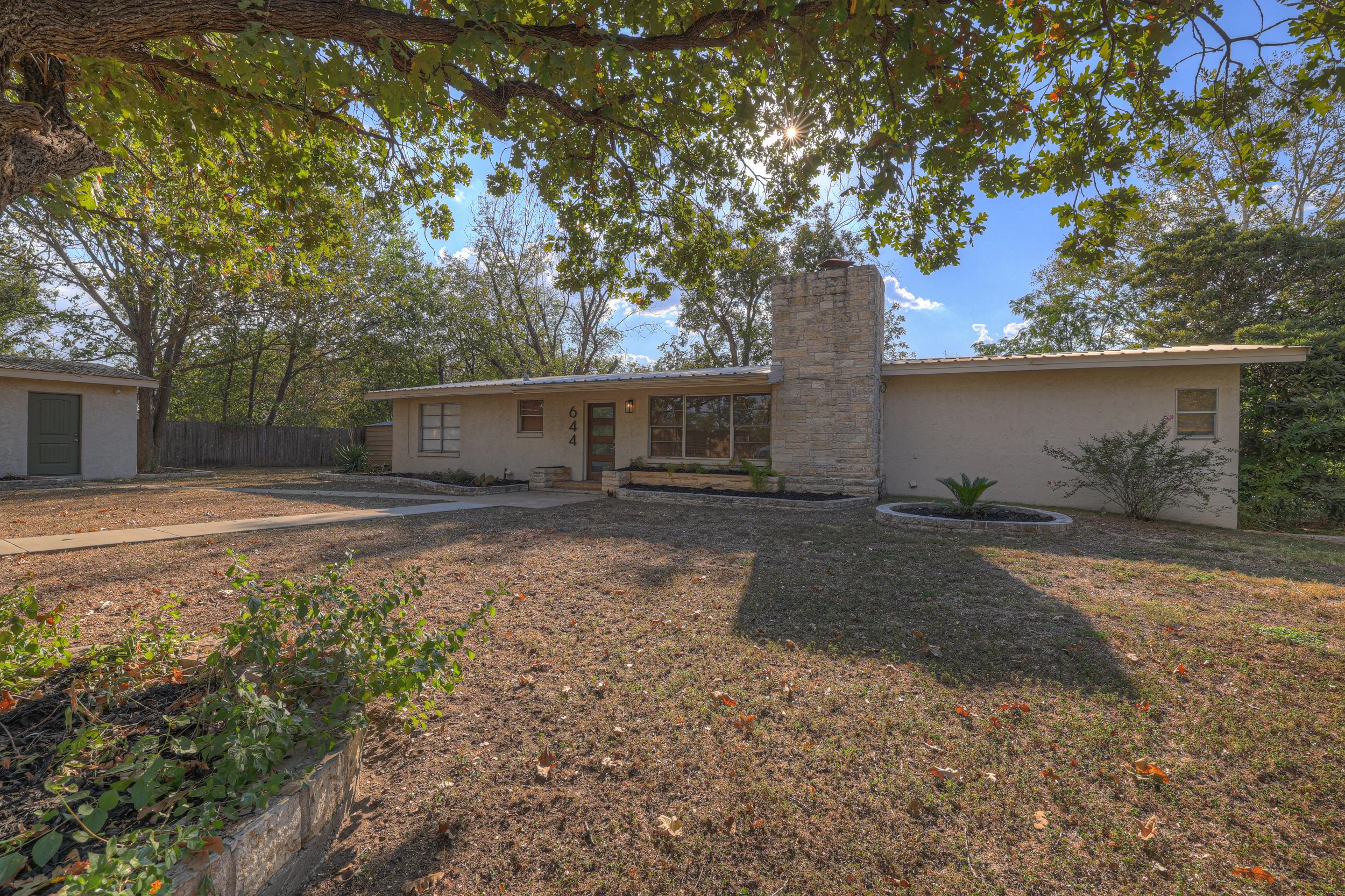 Single-story house with a stone chimney, front yard with sparse grass and a concrete walkway, trees overhead, and blue sky with clouds.