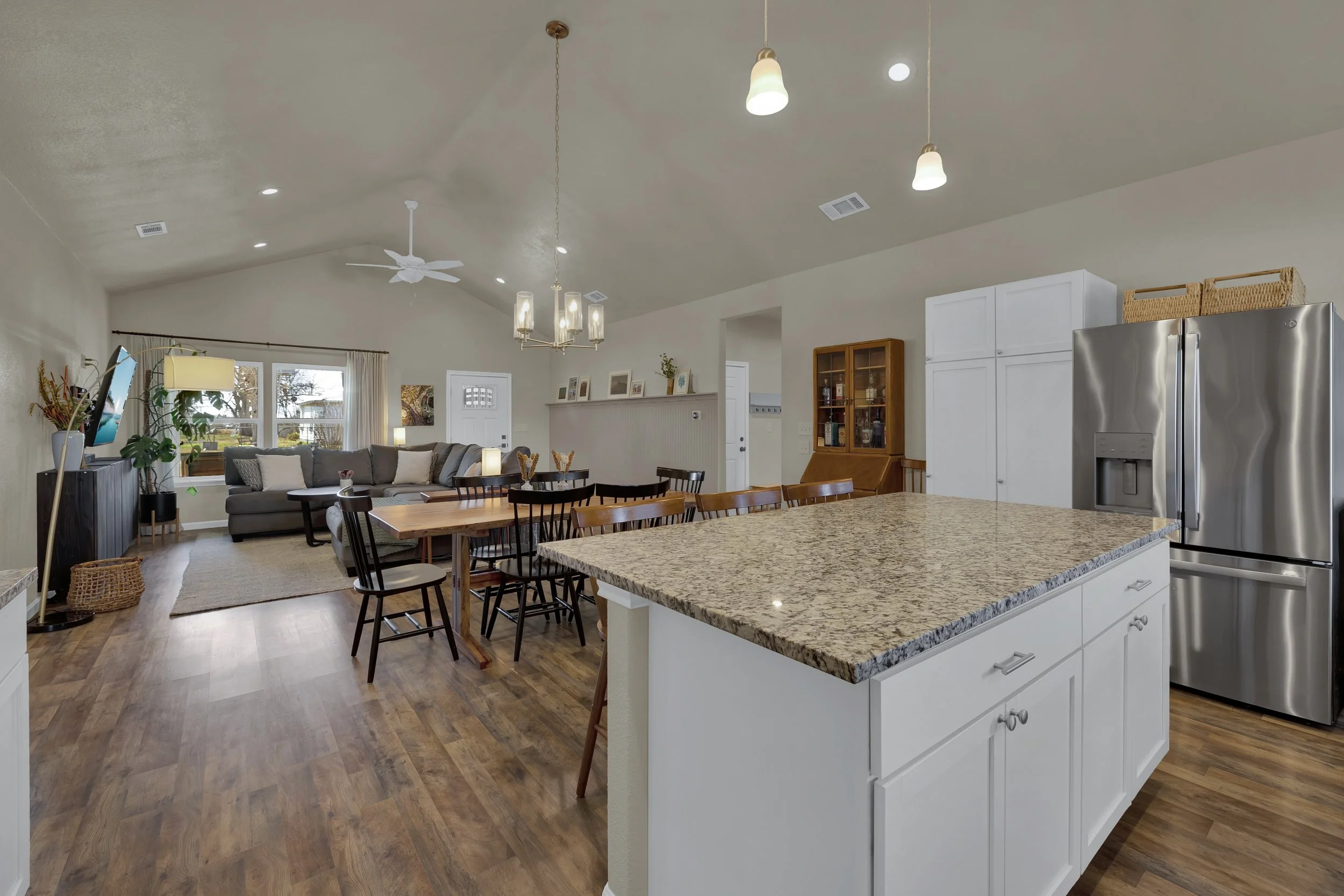 Open-concept kitchen and living room with granite island, white cabinets, stainless steel refrigerator, dining table with black chairs, gray sectional sofa, large window, hardwood floors, and ceiling lights.