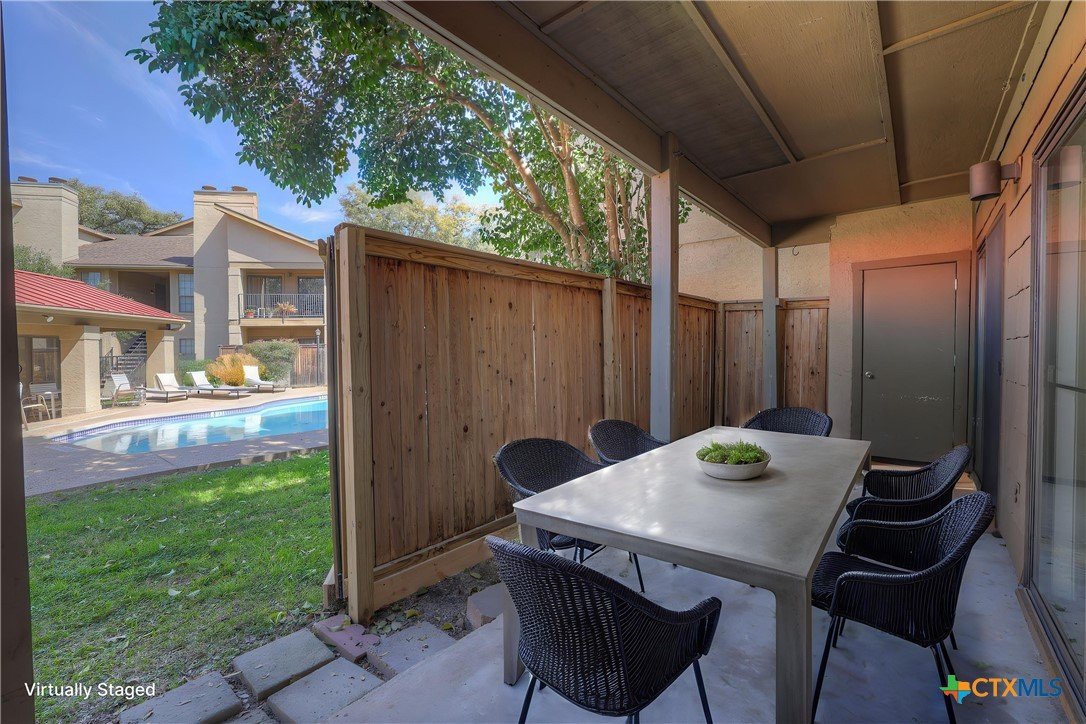 Covered patio area with a white table and six black chairs, surrounded by a wooden privacy fence, overlooking a backyard with a swimming pool and lounge chairs.