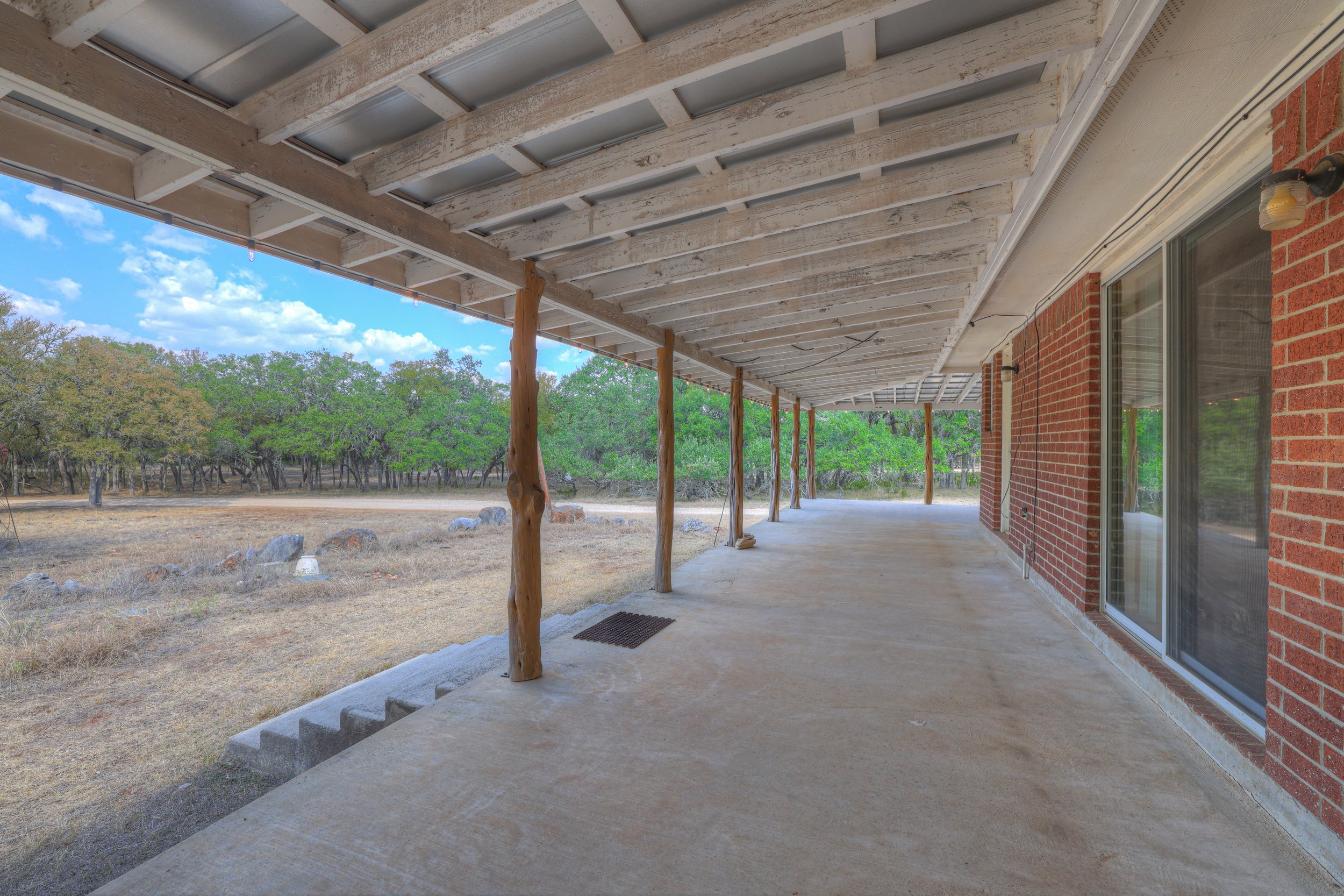 Covered patio with unfinished wood beams, brick house wall with sliding glass door, and scenic outdoor landscape with trees and rocks.