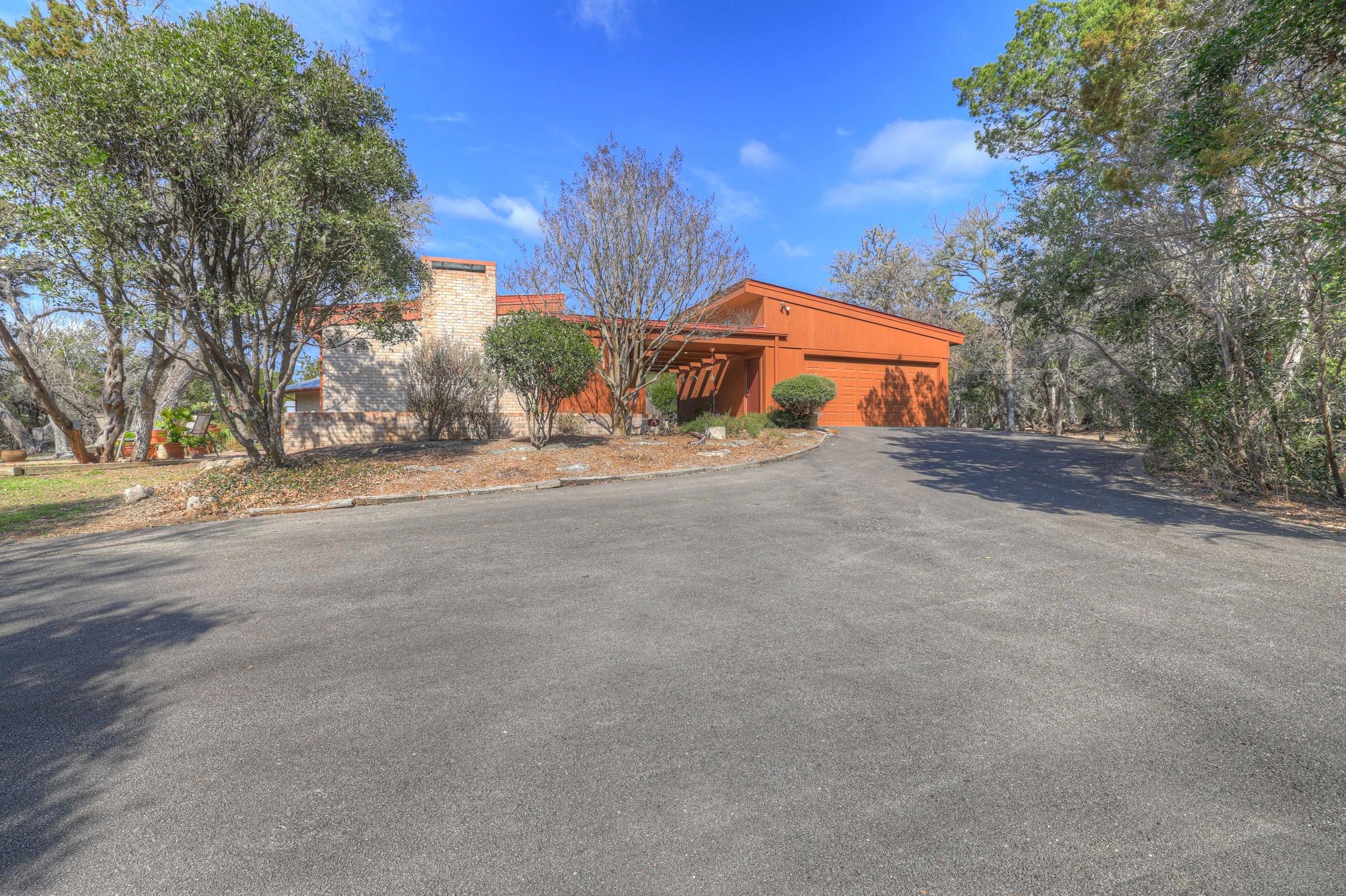 A house with a red exterior, surrounded by trees, with a paved driveway leading up to it under a blue sky.