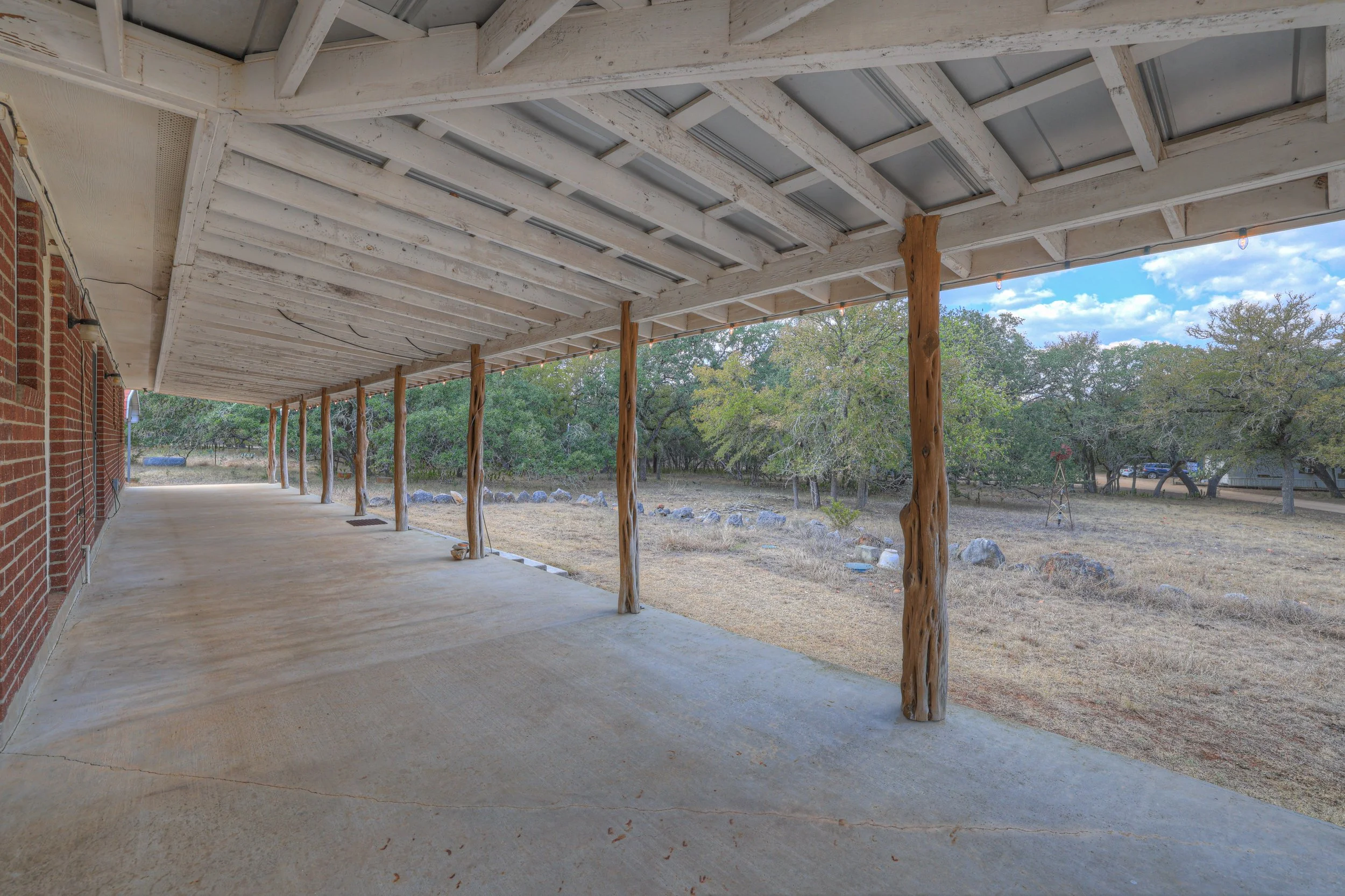 Covered porch with wooden support beams overlooking a yard with trees and rocks.