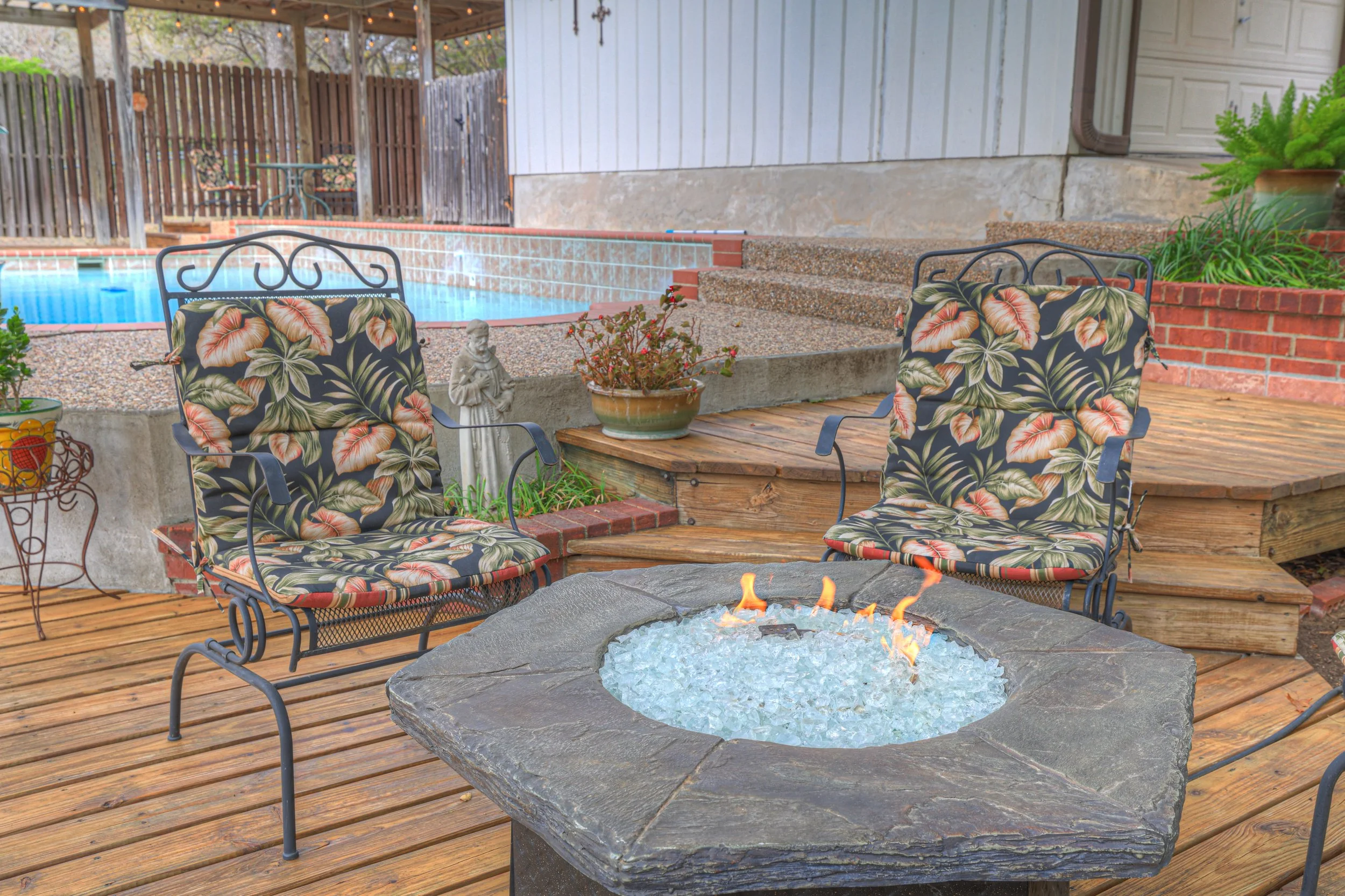 A backyard patio with two metal chairs with floral cushions around a fire pit, in front of a small pool, with potted plants and a statue.