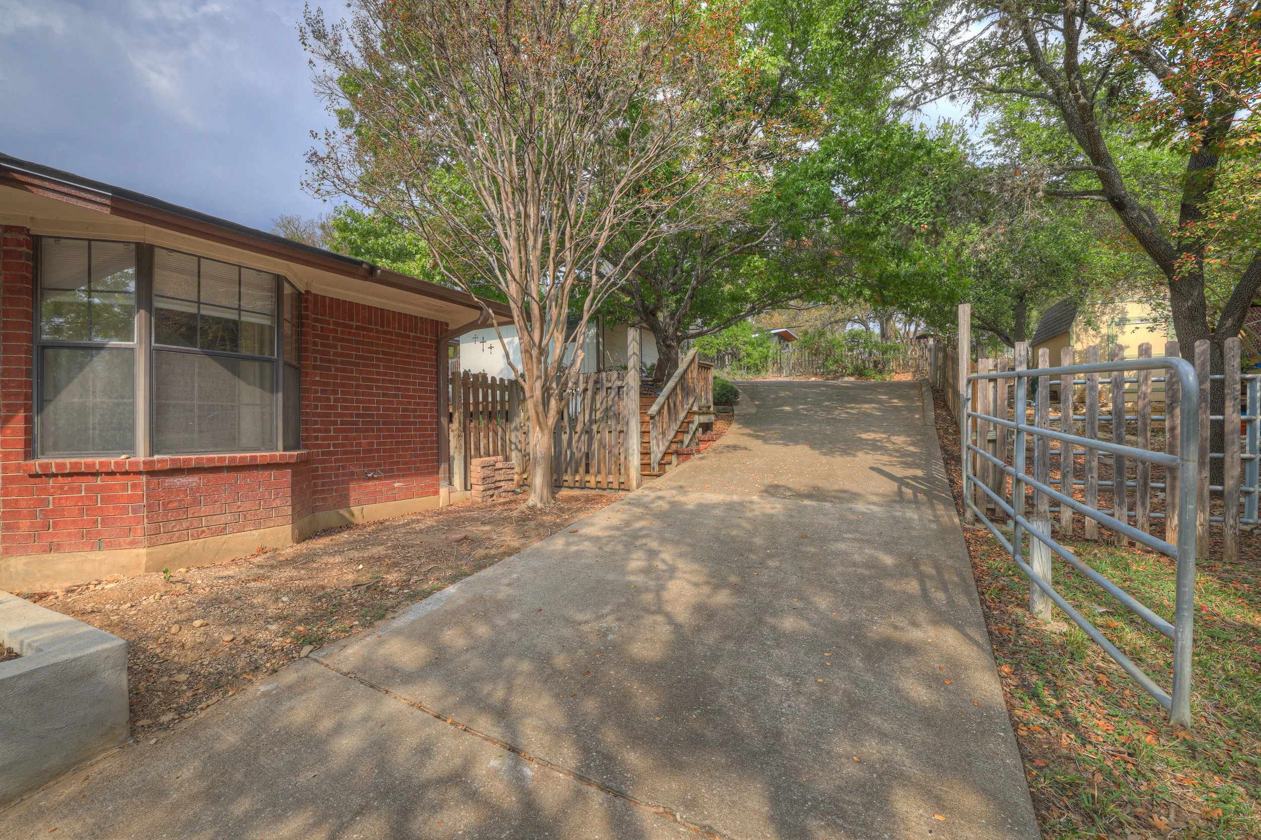 A sloped concrete driveway leading up to a house with a brick exterior and large front window, surrounded by trees and a wooden fence, with shadows cast on the ground.