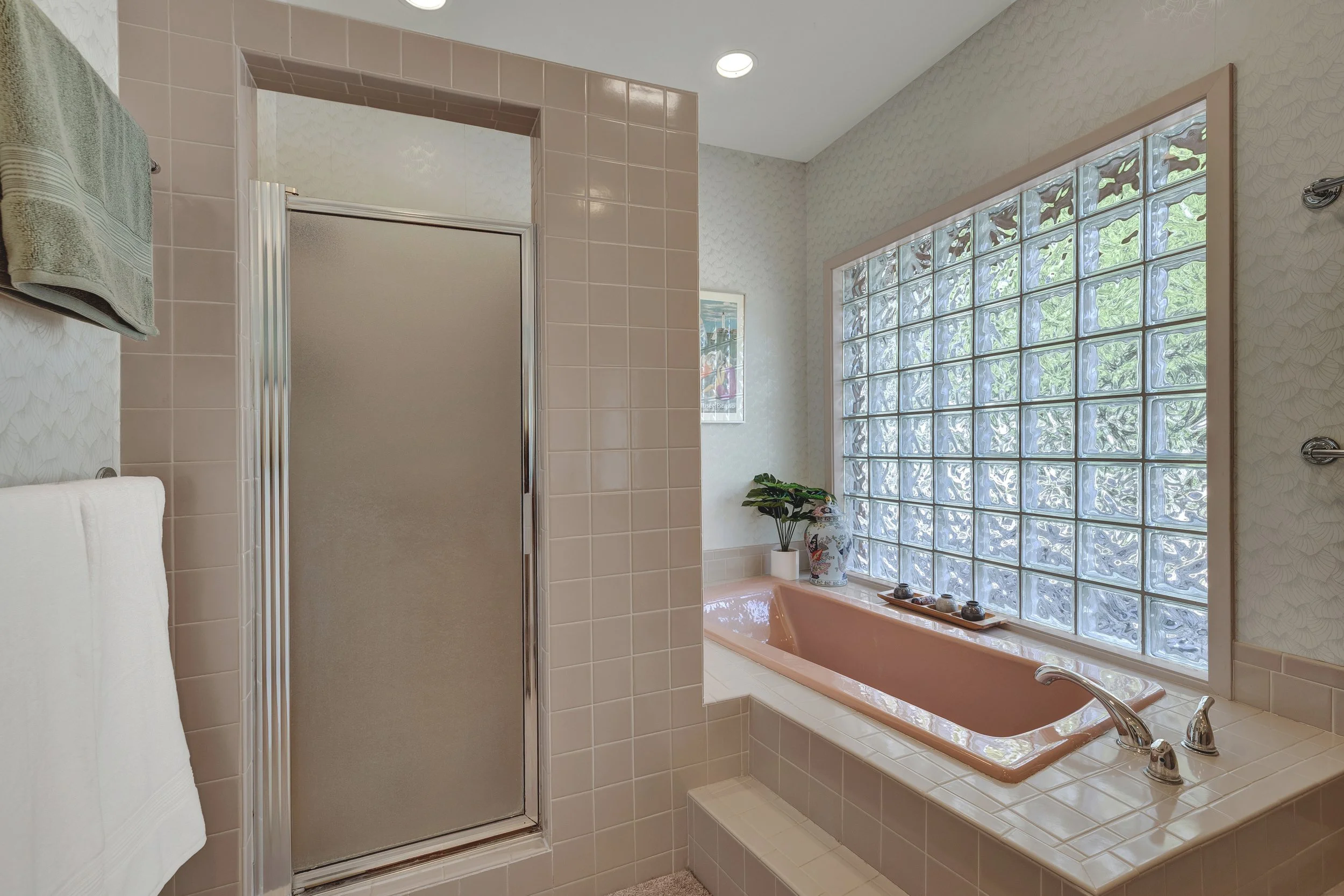 A bathroom featuring a pink bathtub beside a large glass block window, with a potted plant and decorative items on the bathtub edge, and a frosted glass shower door to the left.