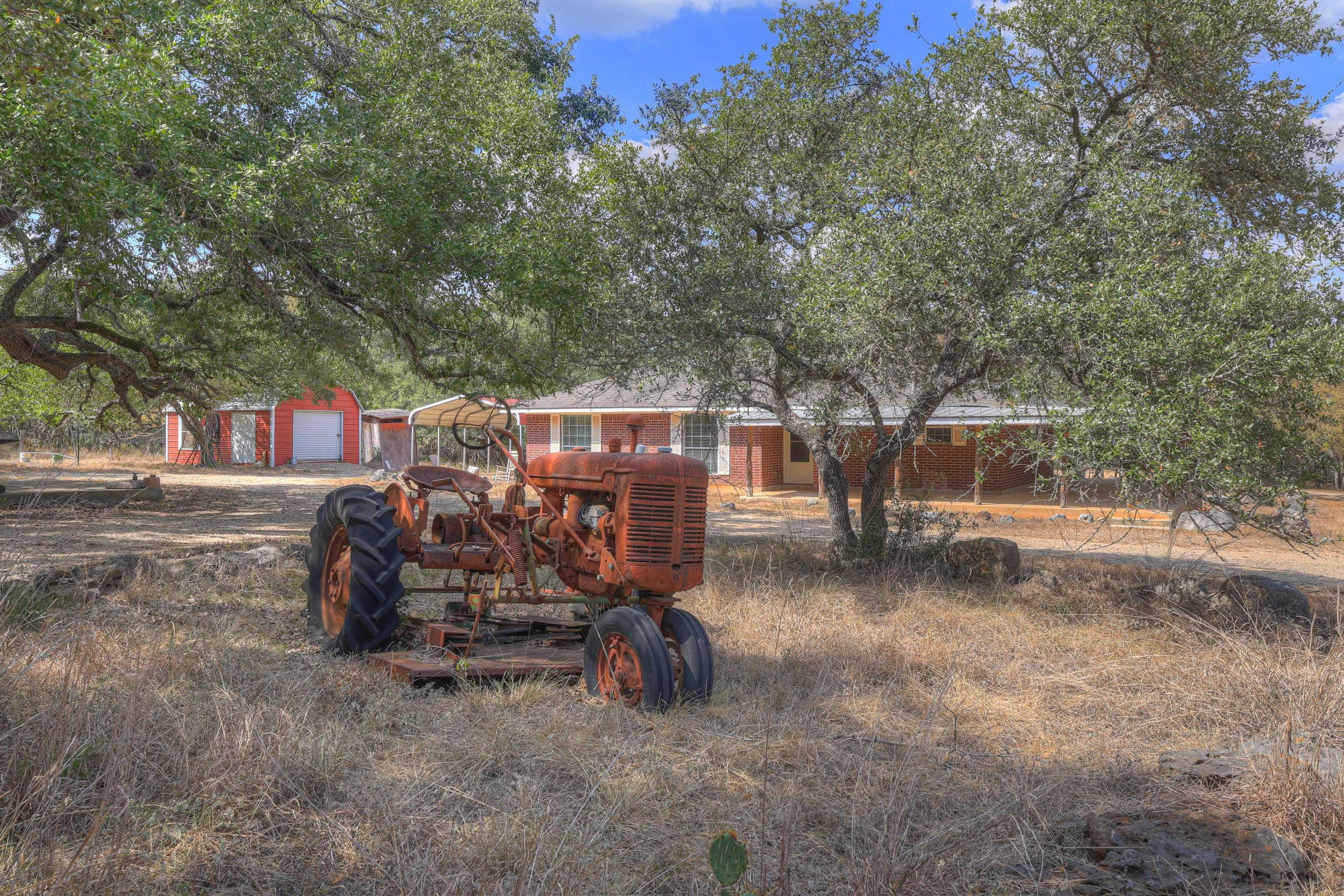 A rusty vintage tractor on dry grass in front of a large tree, with a brick house and a red shed in the background under a partly cloudy sky.