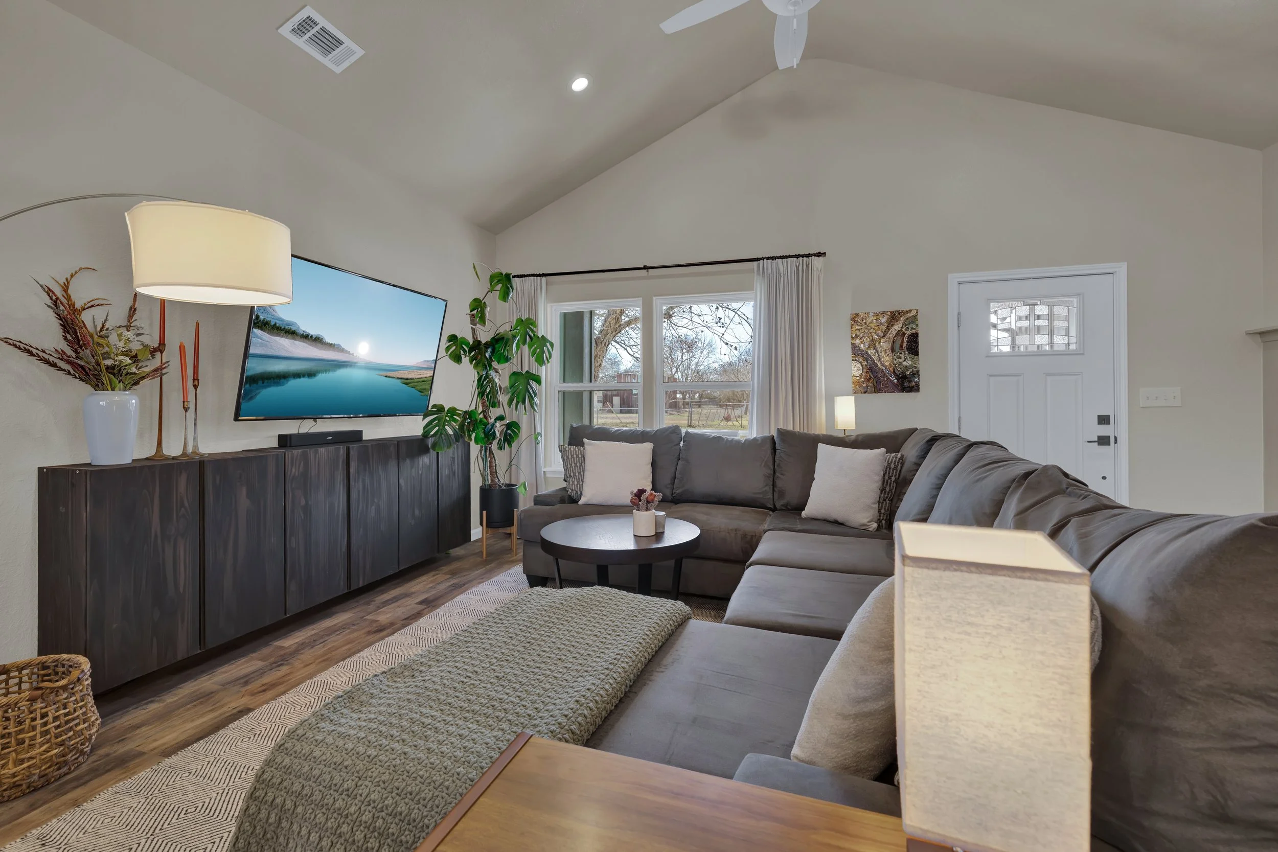 Living room with a large gray sectional sofa, a round coffee table, a tall potted plant, and a large wall-mounted television. There is a window with white curtains behind the sofa, and the front door is visible on the right side. The room has a high,