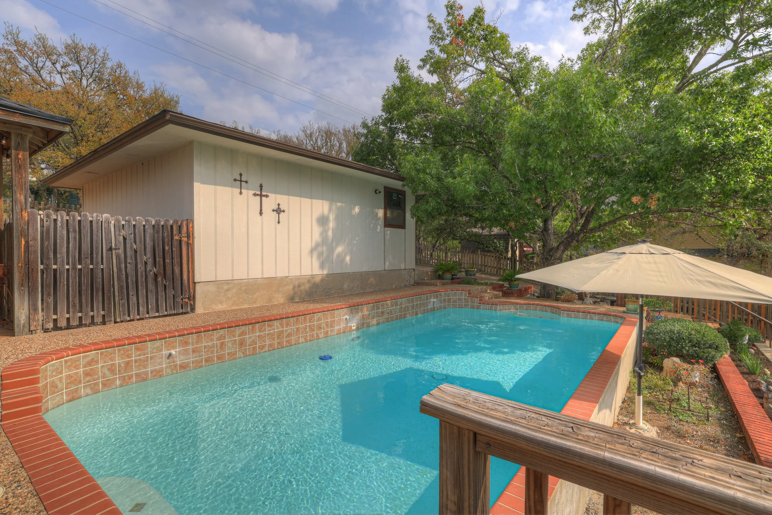 A backyard with a swimming pool, a large tree, a white building with crosses on the wall, and an outdoor umbrella.