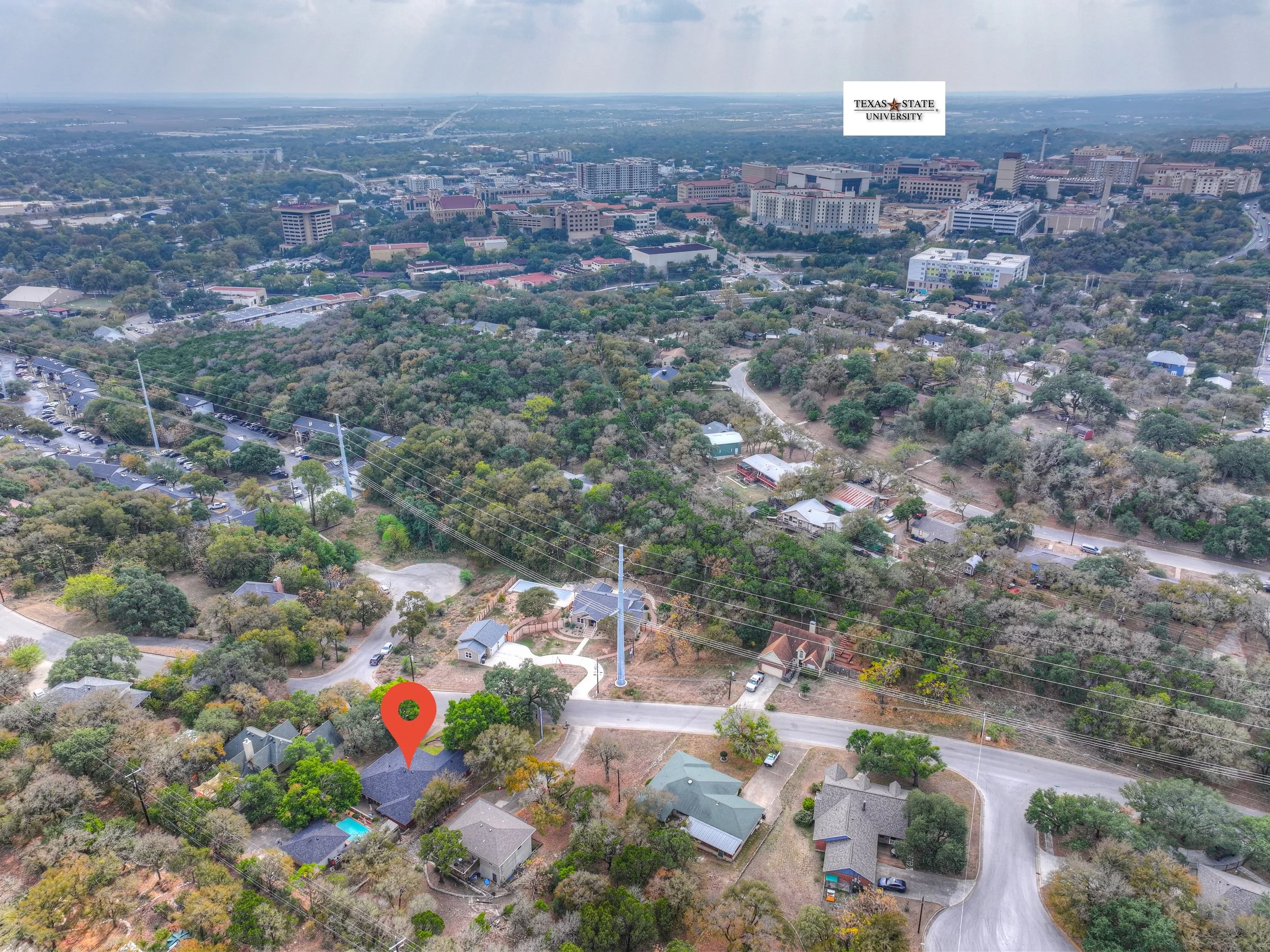 An aerial view of a residential neighborhood with houses, trees, and roads, with Texas State University buildings visible in the background and a label pointing to a specific house.