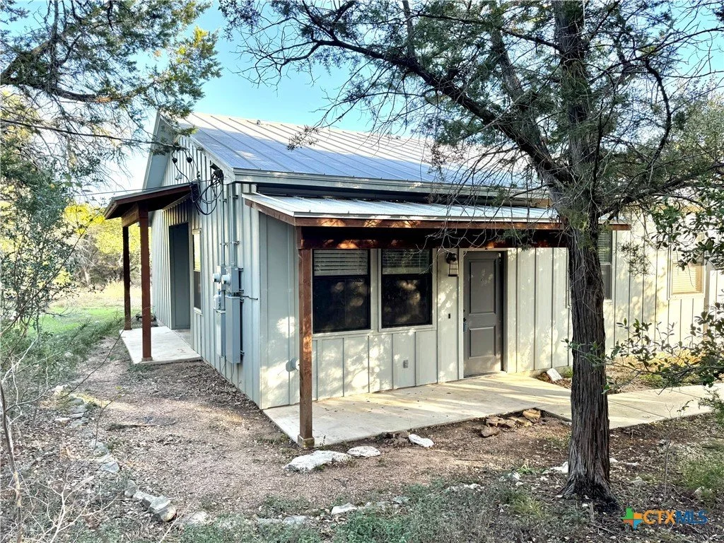 Small metal building with a metal roof, a small covered porch, and a door on the front, surrounded by trees and dirt ground.