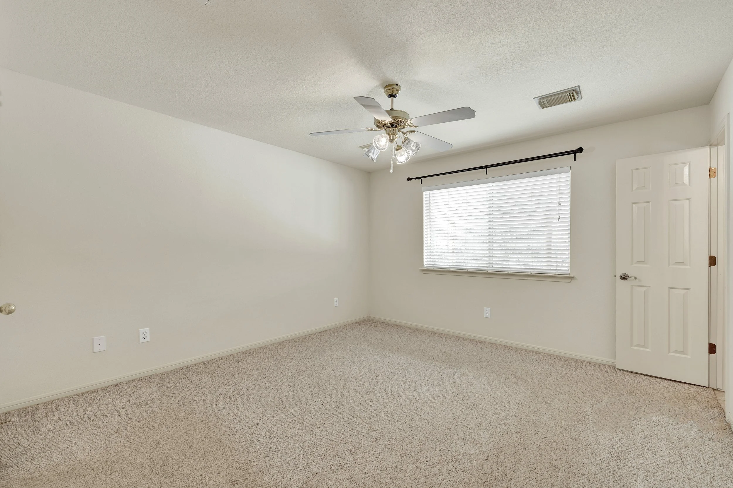 Empty room with beige carpet, white walls, a window with blinds, a ceiling fan with lights, and a closed door.