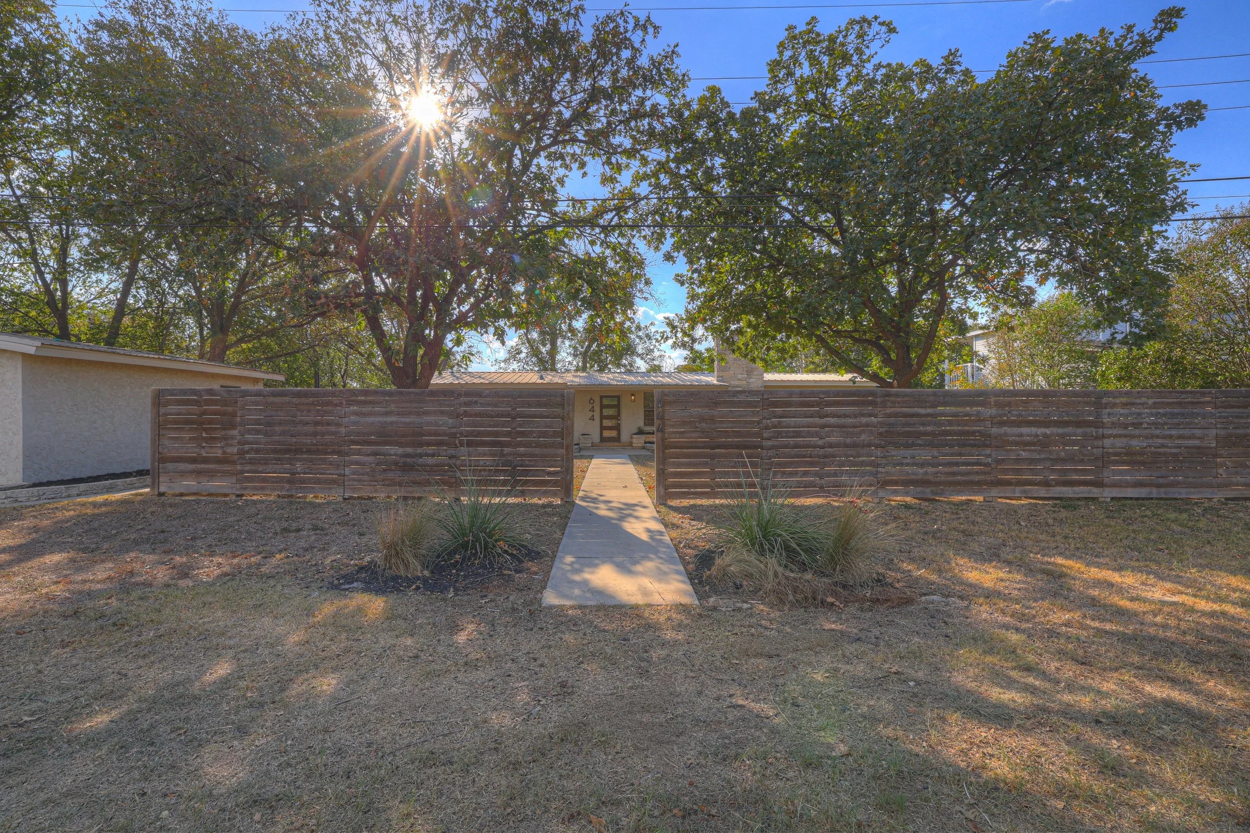 Front yard with a concrete pathway leading to a house entrance, surrounded by a wooden fence, grass, and two large trees with the sun shining through the branches.