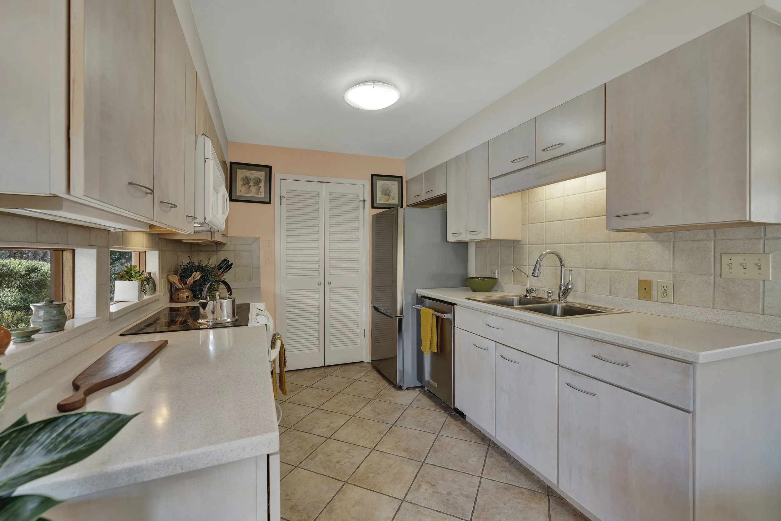 Clean kitchen with white cabinets, beige tiled floor, double sink, refrigerator, stove, and small windows with greenery outside.