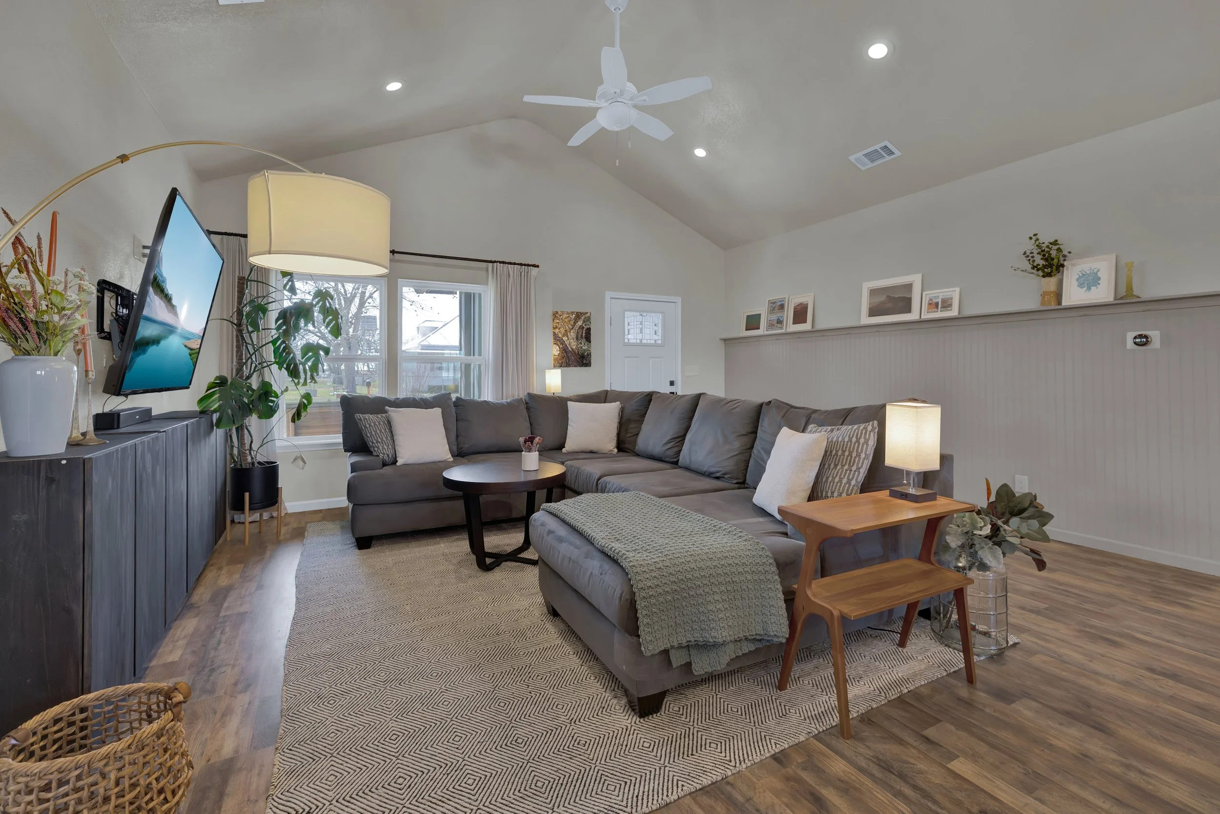 Living room with a large gray sectional sofa, wooden side table with lamp, coffee table with plant, TV mounted on dark wood cabinet, window with white curtains, and ceiling fan with lights.
