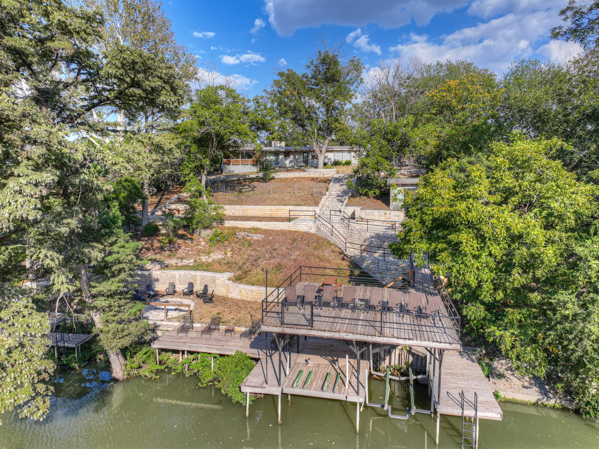 Aerial view of a riverfront property with multiple wooden decks, outdoor chairs, and trees, under a partly cloudy sky.