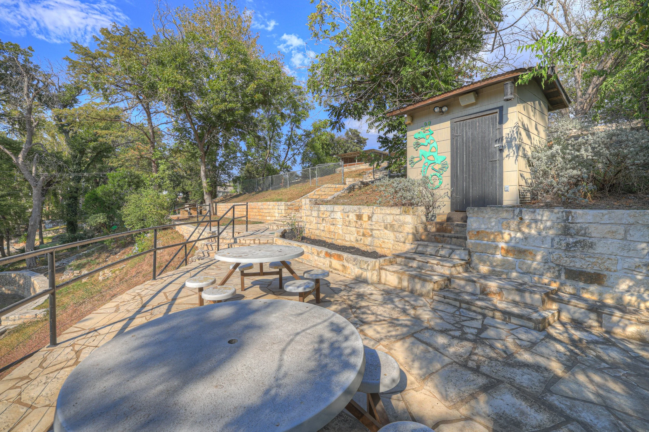 Outdoor patio area with stone pathways, round concrete tables with attached stools, surrounded by trees and bushes, with a small beige building featuring a painted lizard on the side and steps leading up to it.
