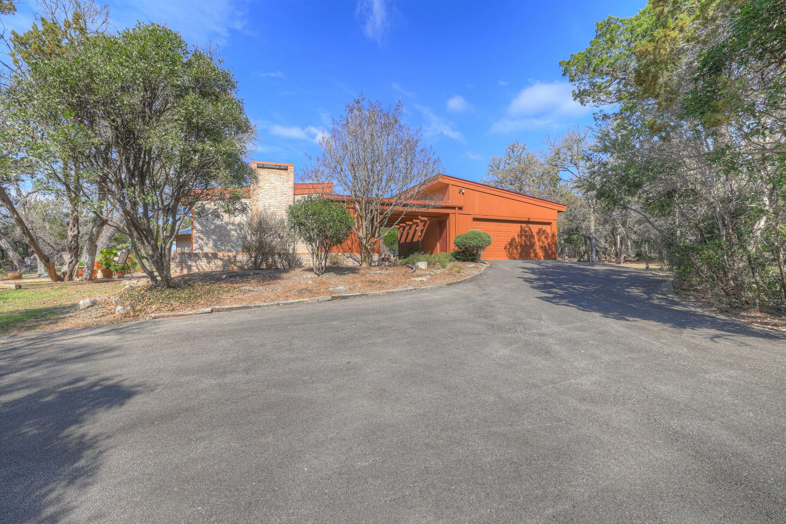 A house with a red garage door, surrounded by trees and bushes, with a curved asphalt driveway leading to the garage, under a partly cloudy blue sky.