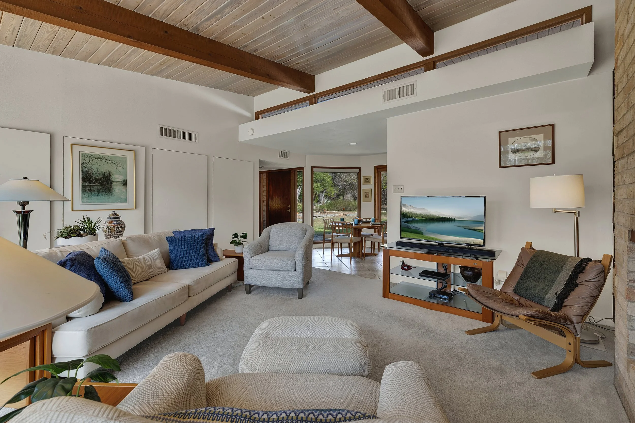 A living room with white walls, a beige sofa with blue and beige pillows, a gray armchair, a wooden TV stand with a flat-screen TV, a brown leather lounge chair with a throw blanket, a floor lamp, and framed artwork on the walls. The room has a woode