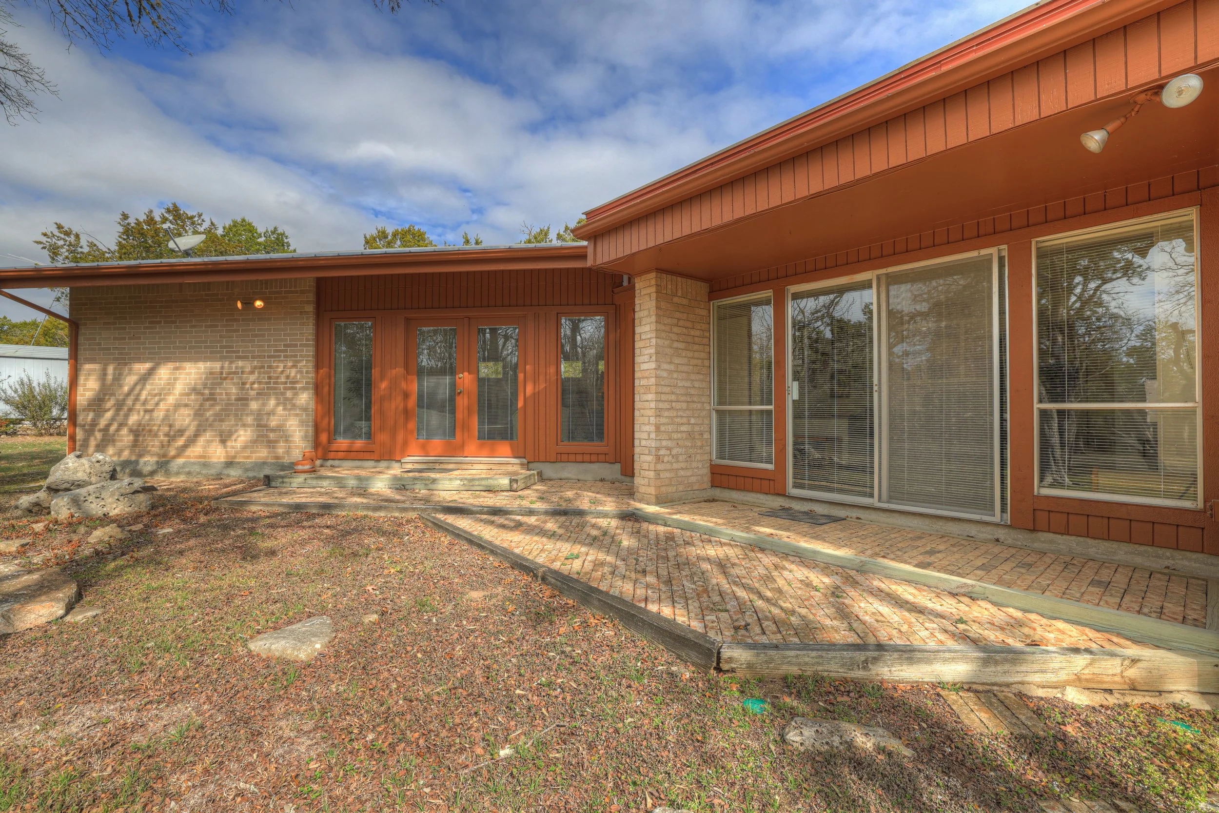 Backyard view of a single-story brick house with large windows and a sliding glass door, a small set of stairs leading to the door, brick patio area, and a wood and brick exterior, under a partly cloudy sky.