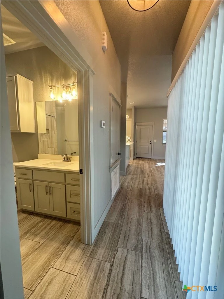 View of a hallway with a bathroom on the left and a living area at the end, featuring tile flooring, light-colored walls, and vertical blinds on the right window.