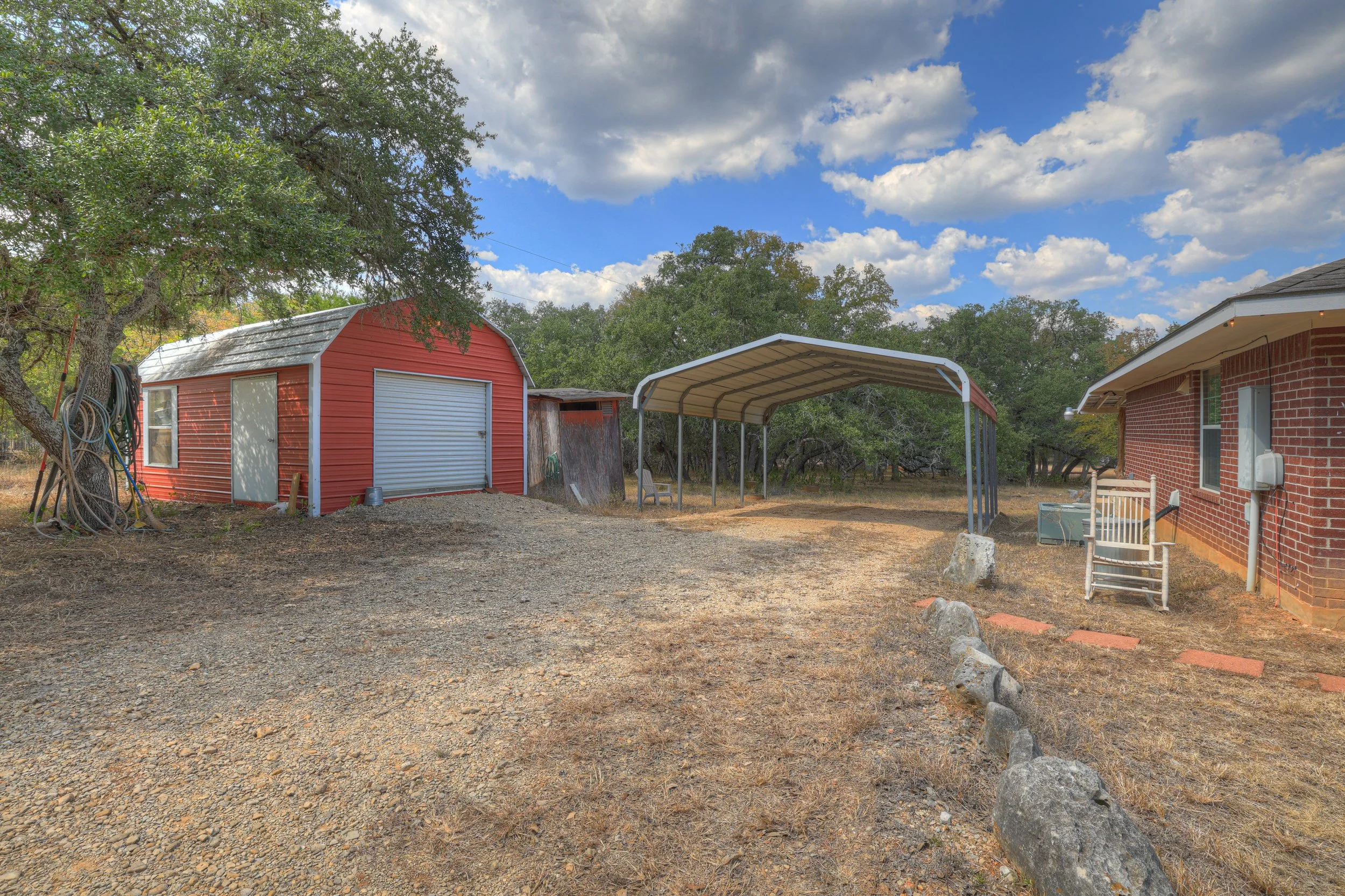 A backyard with a red shed with a closed roll-up door, a small rusted shed, a carport with a curved roof, a brick house, a wooded background, and a gravel yard under a partly cloudy sky.