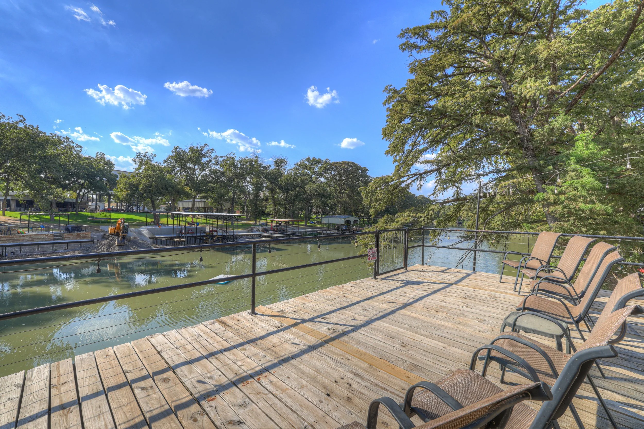 Wooden deck with chairs overlooking a river, with trees and a construction site in the background on a sunny day.
