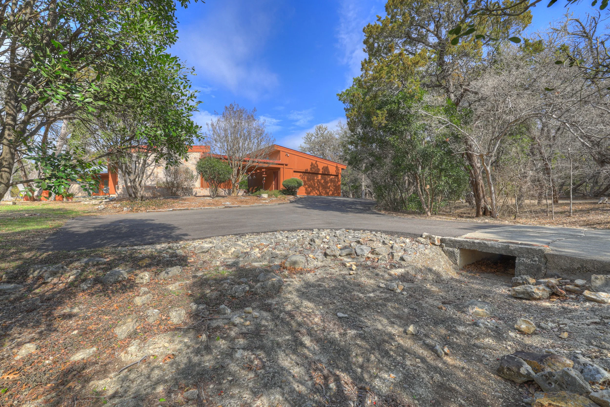 Residential house with a brown exterior surrounded by trees, a paved driveway, and a small bridge crossing a dry creek bed in front.