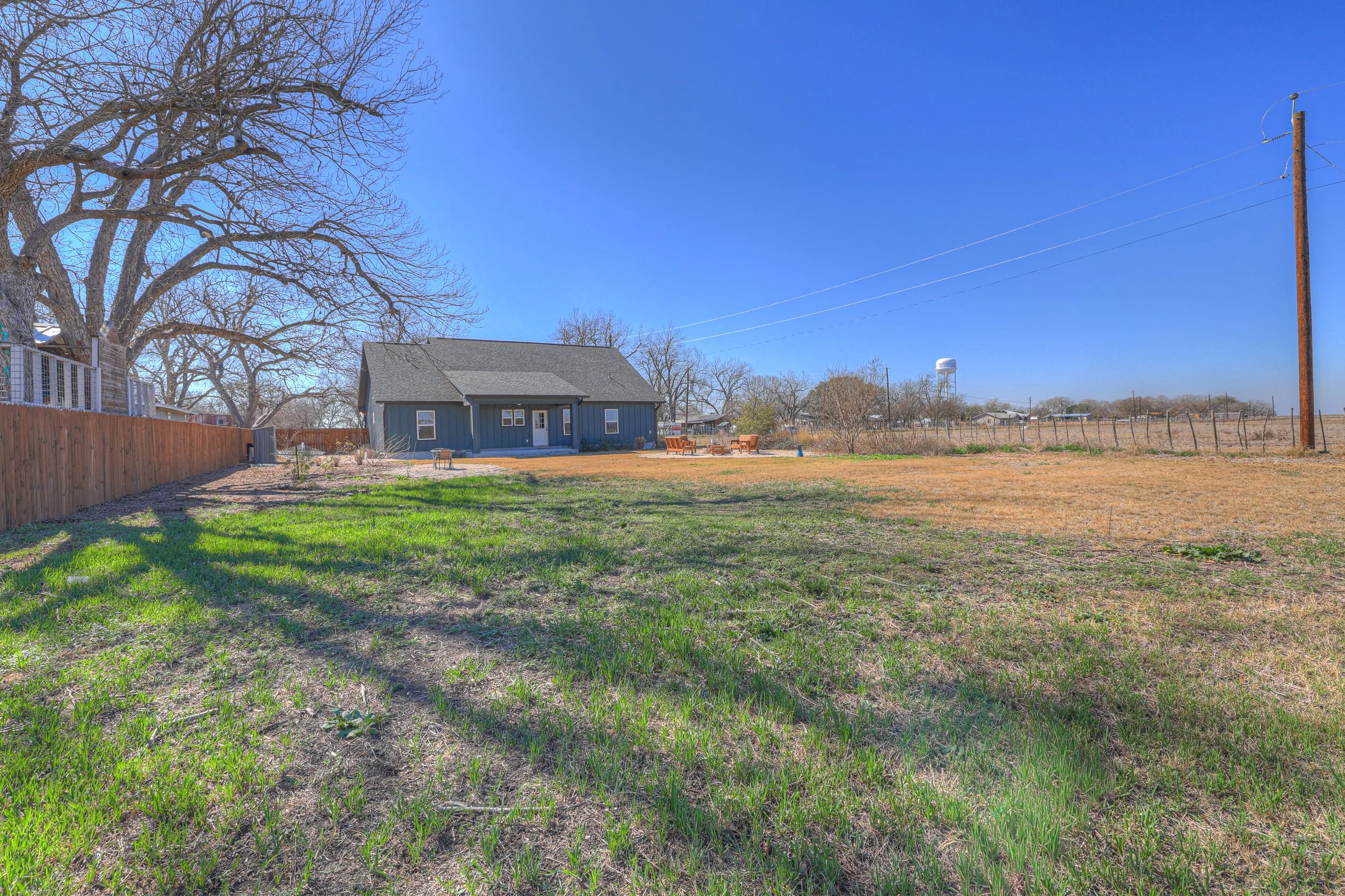 A backyard with a mix of green and brown grass, a gray house with a porch, a large leafless tree, a wooden fence on the left, and a utility pole on the right against a clear blue sky.