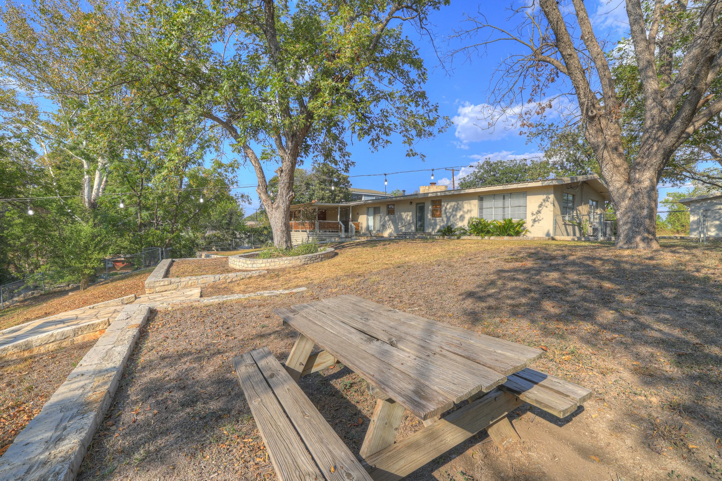 Outdoor backyard area with a wooden picnic table, large trees, and a single-story house in the background under a blue sky.