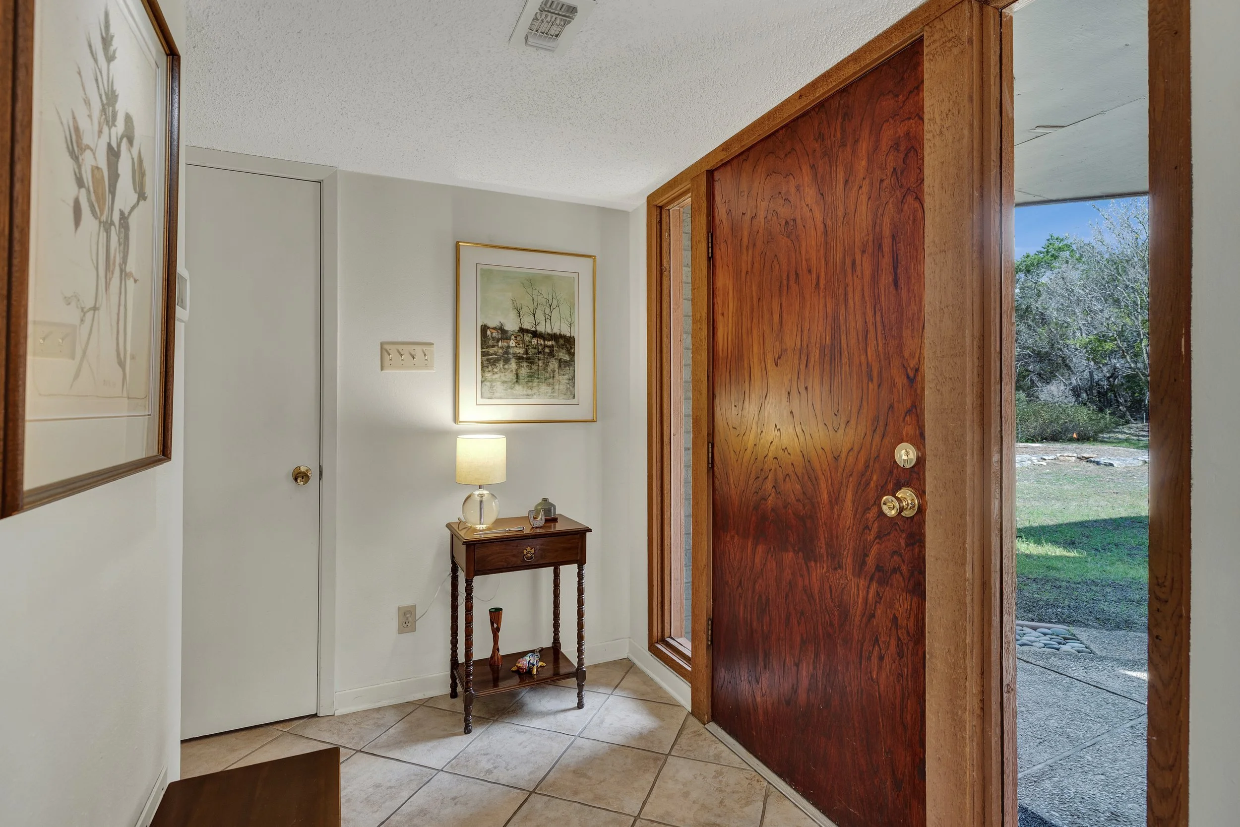 Entryway with tiled floor, open front door showing outside yard, small side table with a lamp and decorations, framed pictures on the white wall.