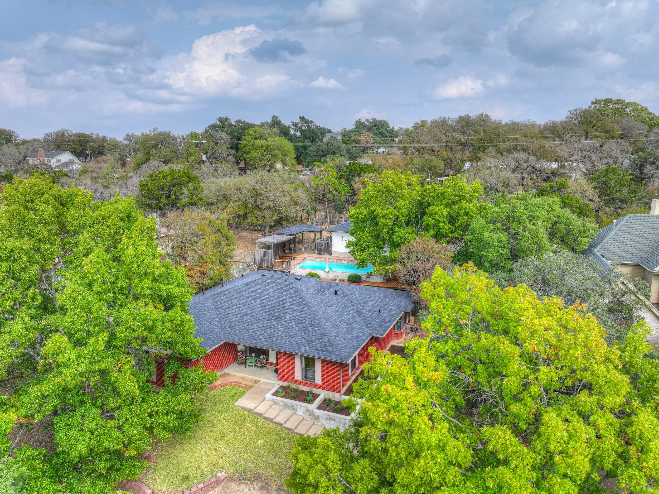 Aerial view of a house with a dark gray roof, surrounded by green trees, with a backyard featuring a swimming pool and a wooden fence, under a partly cloudy sky.