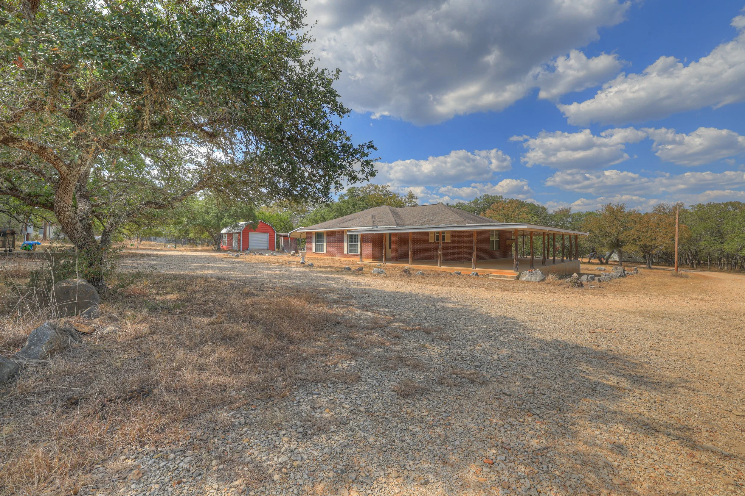 A house with a porch sits on a dry, rocky yard under a partly cloudy sky. There is a red shed in the background and a large tree on the left side of the image.