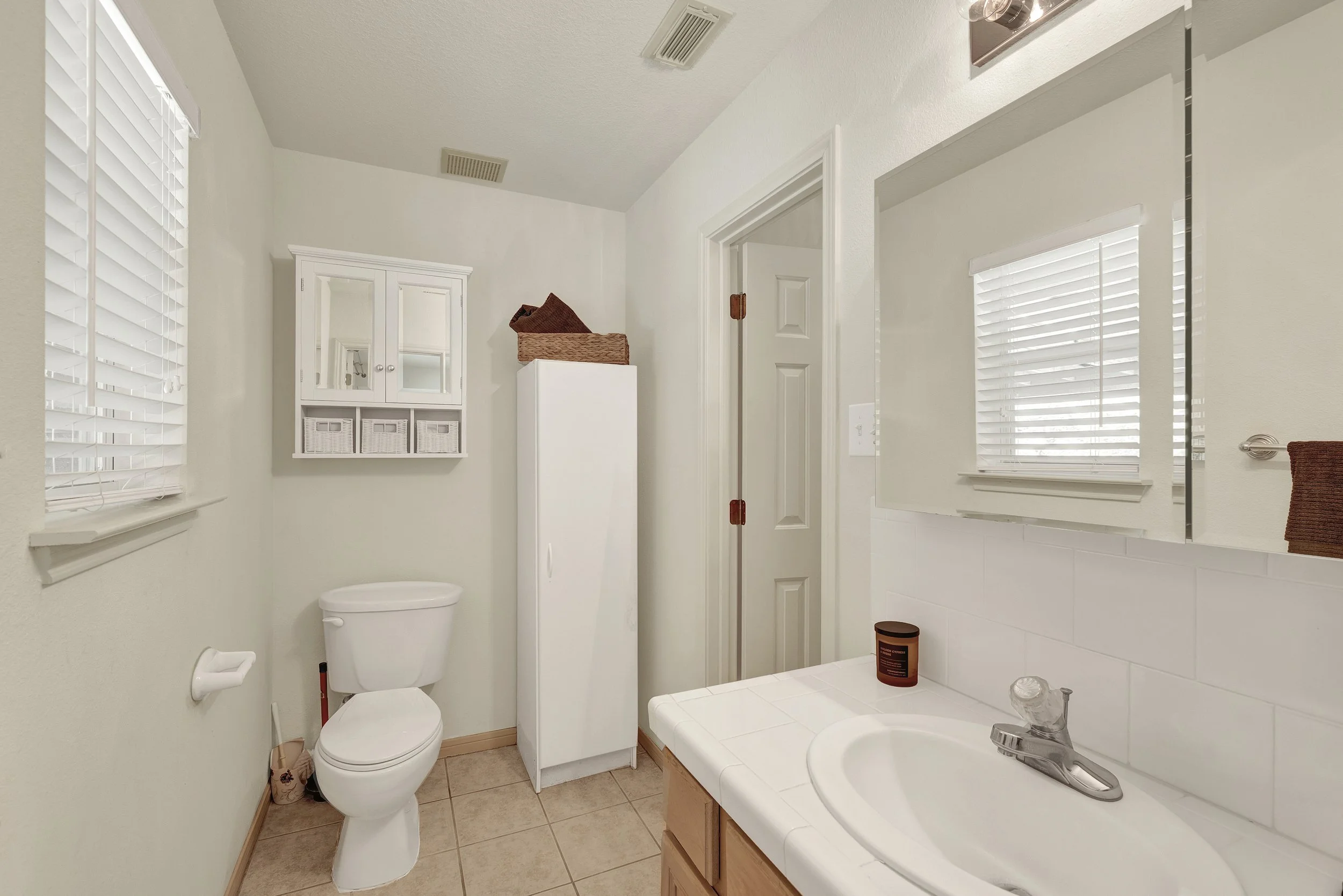 A small bathroom with a white toilet, a white storage cabinet, a tiled sink with a mirror above, brown towels, and beige tiled flooring.