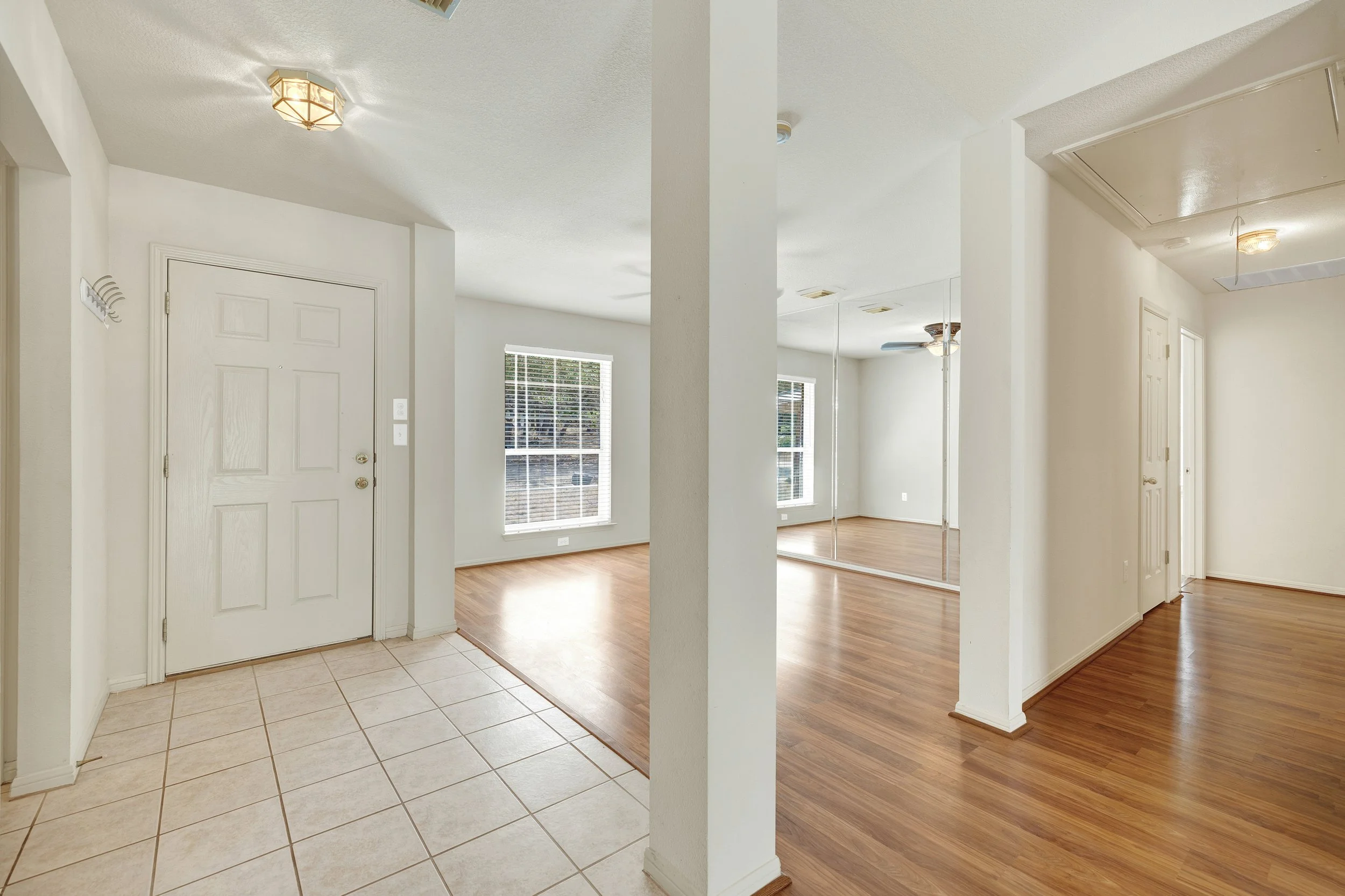 Empty interior of a house with an entryway featuring tiled floor, white walls, and a white front door; adjacent room has large windows, wood flooring, and mirrored closet doors.