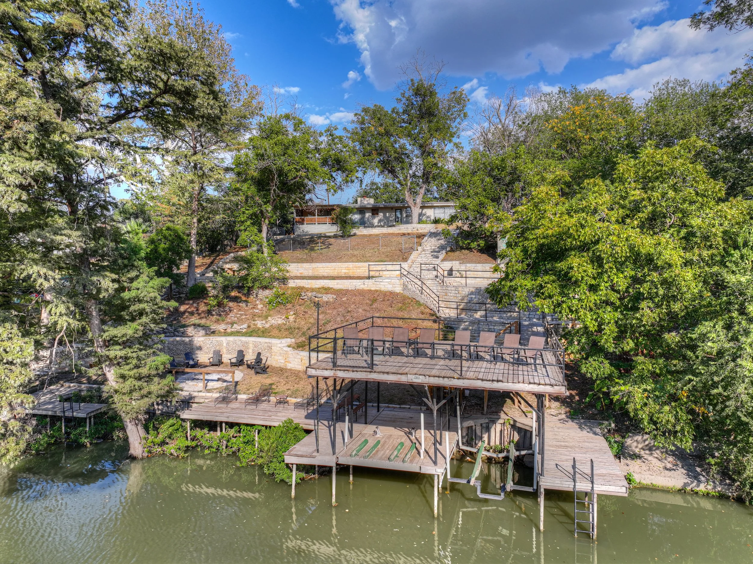 A backyard with trees, stairs, and a deck overlooking a body of water. The deck has outdoor chairs and a table, with stairs leading up to a house on a hill.