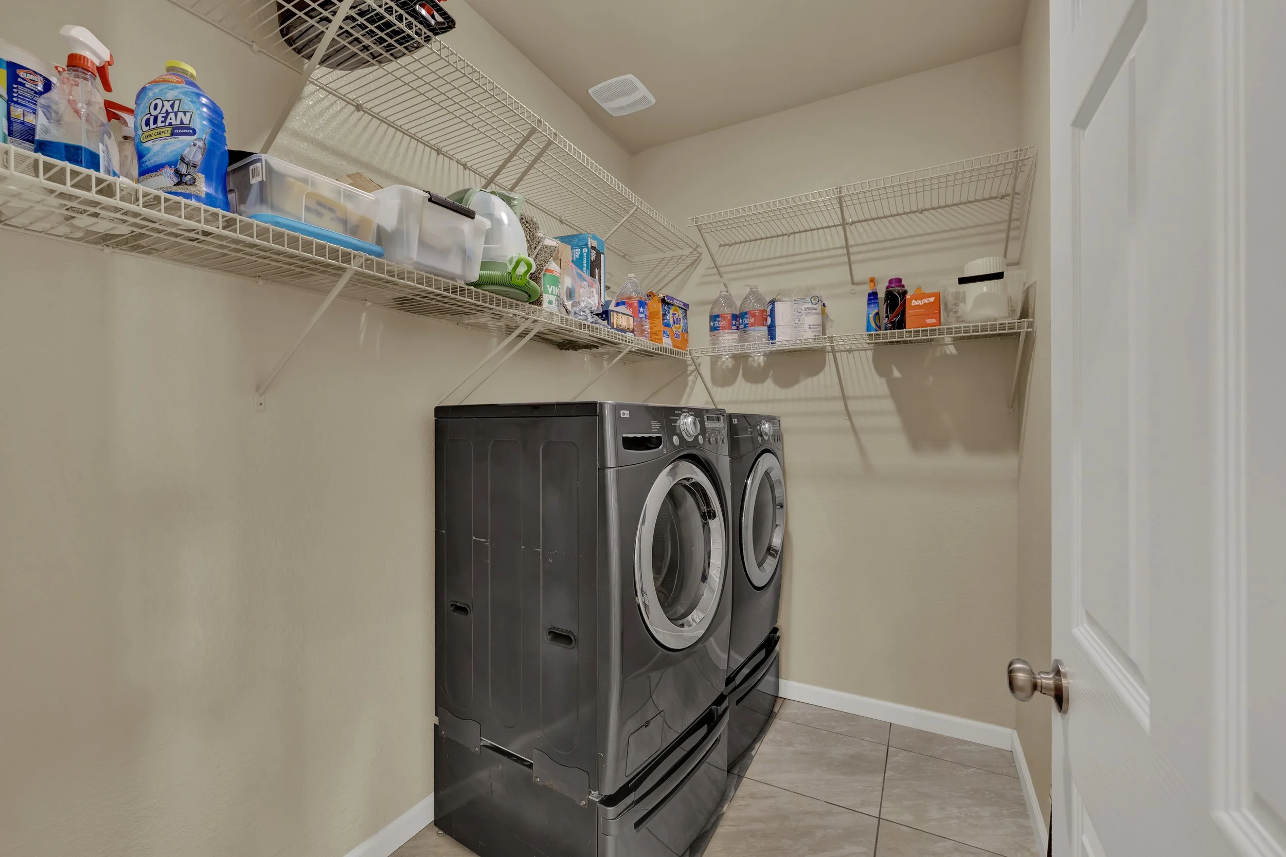 A laundry room with a front-loading washer and dryer, and wire shelves holding cleaning supplies, detergent bottles, water bottles, and boxes.