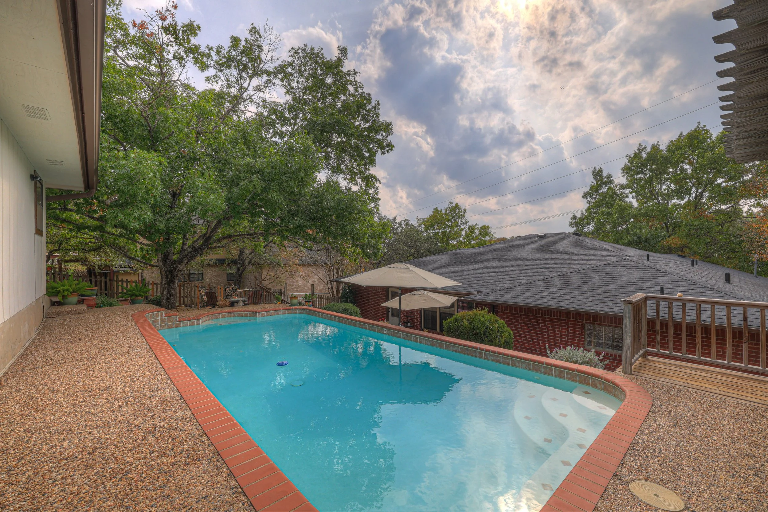 Backyard with swimming pool, large trees, and neighboring houses with brick walls and shaded patios under cloudy sky.