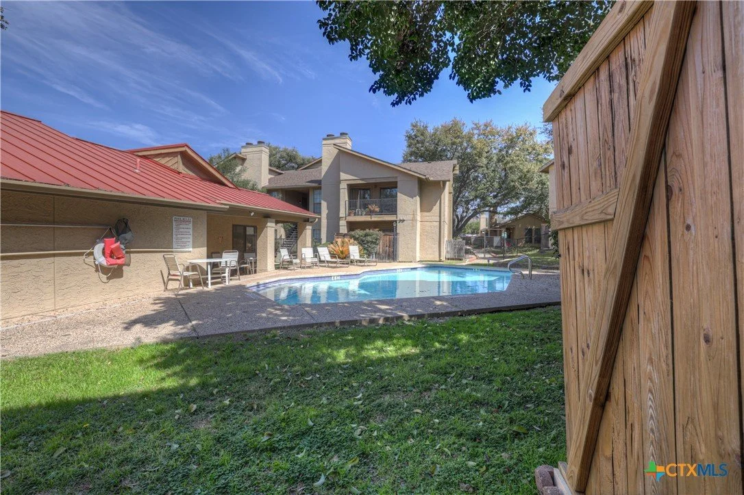 Apartment complex pool area with lounge chairs, a table, and a wooden shed on grass, under a blue sky with some clouds.