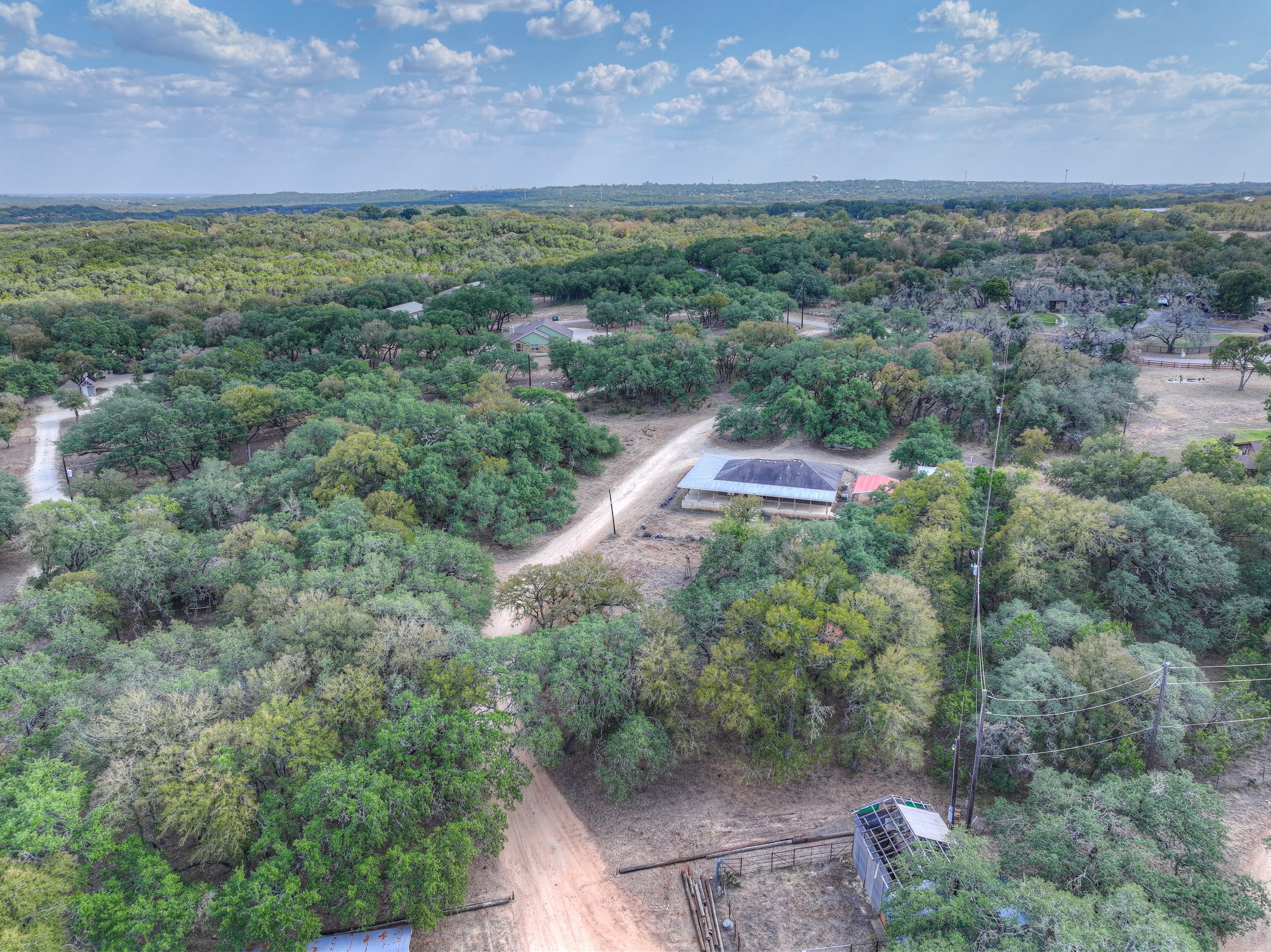 Aerial view of a rural area with trees, dirt roads, and some buildings, including a house with a metal roof and a small greenhouse.