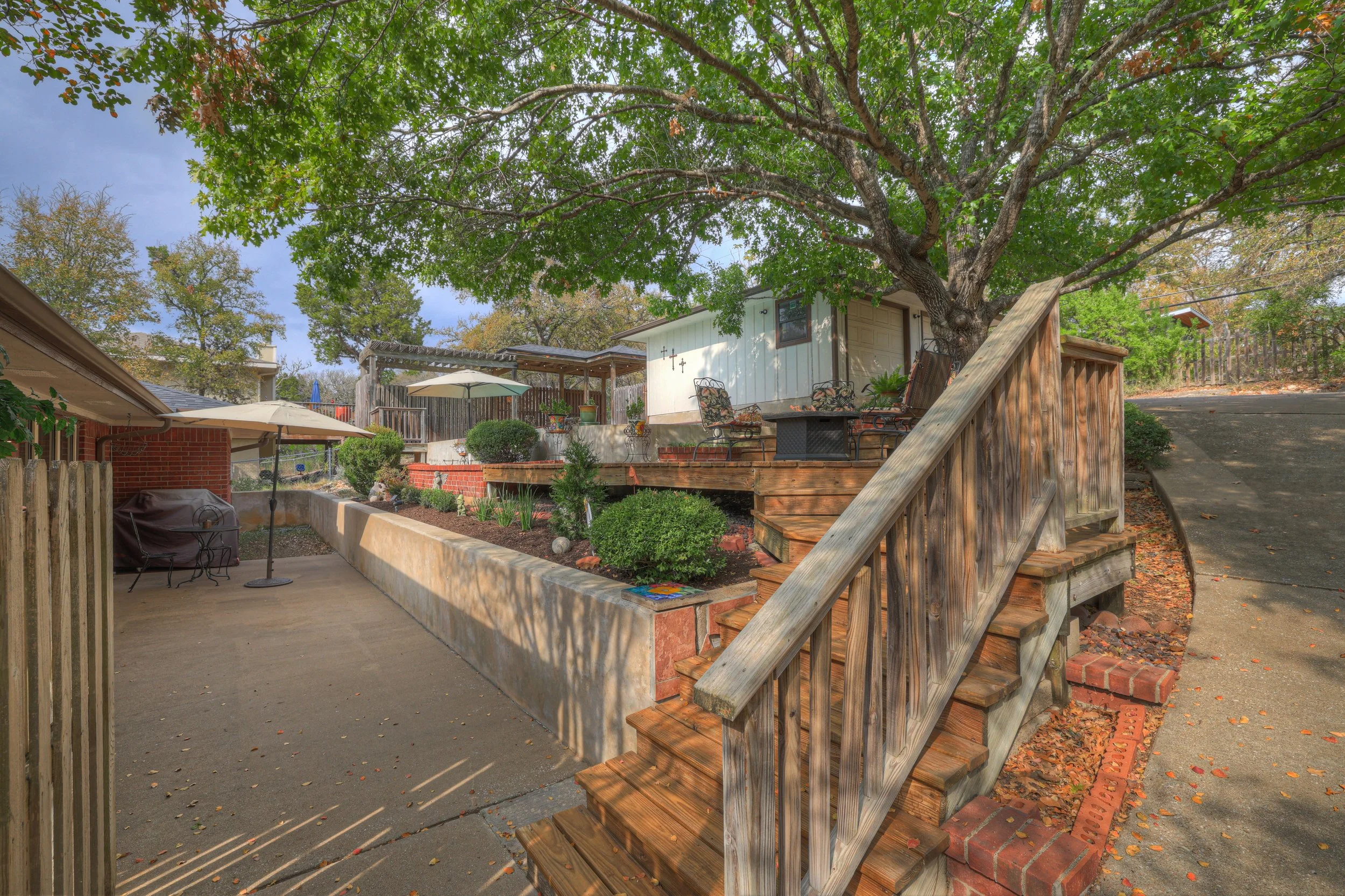 A backyard patio with wooden stairs and railing, outdoor furniture including chairs and an umbrella, a garden area with various plants, a large tree providing shade, and a small building in the background, on a sunny day.