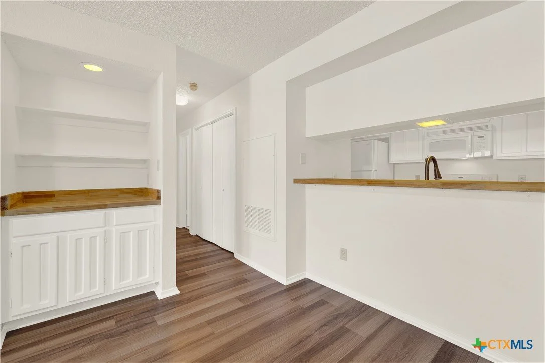 An open kitchen with white cabinets, a wooden countertop, and a pass-through window to a dining area with wooden flooring.
