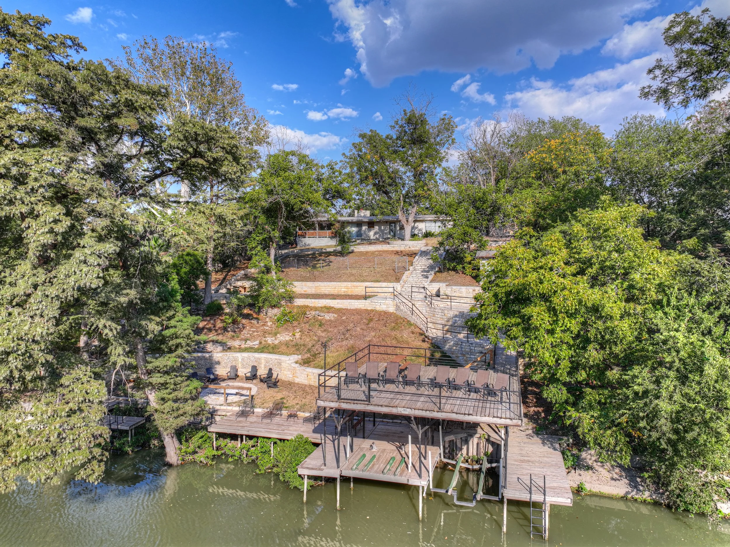 A multi-level house on a hillside surrounded by trees, featuring a deck with outdoor chairs and a dock extending over a body of water.