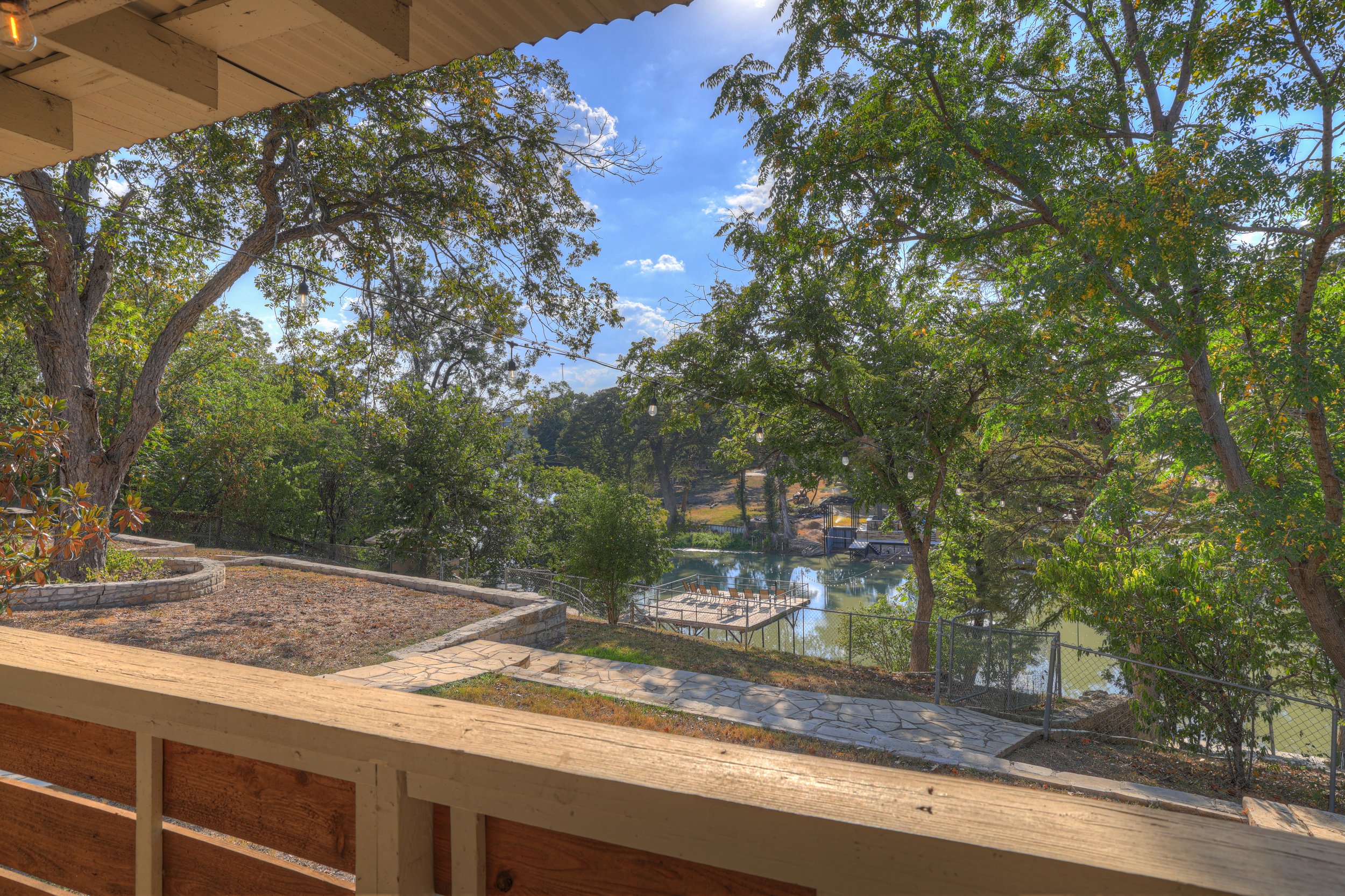 View from a porch overlooking a backyard with trees, a pond, a small dock with chairs, and a stone pathway, under a partly cloudy sky.