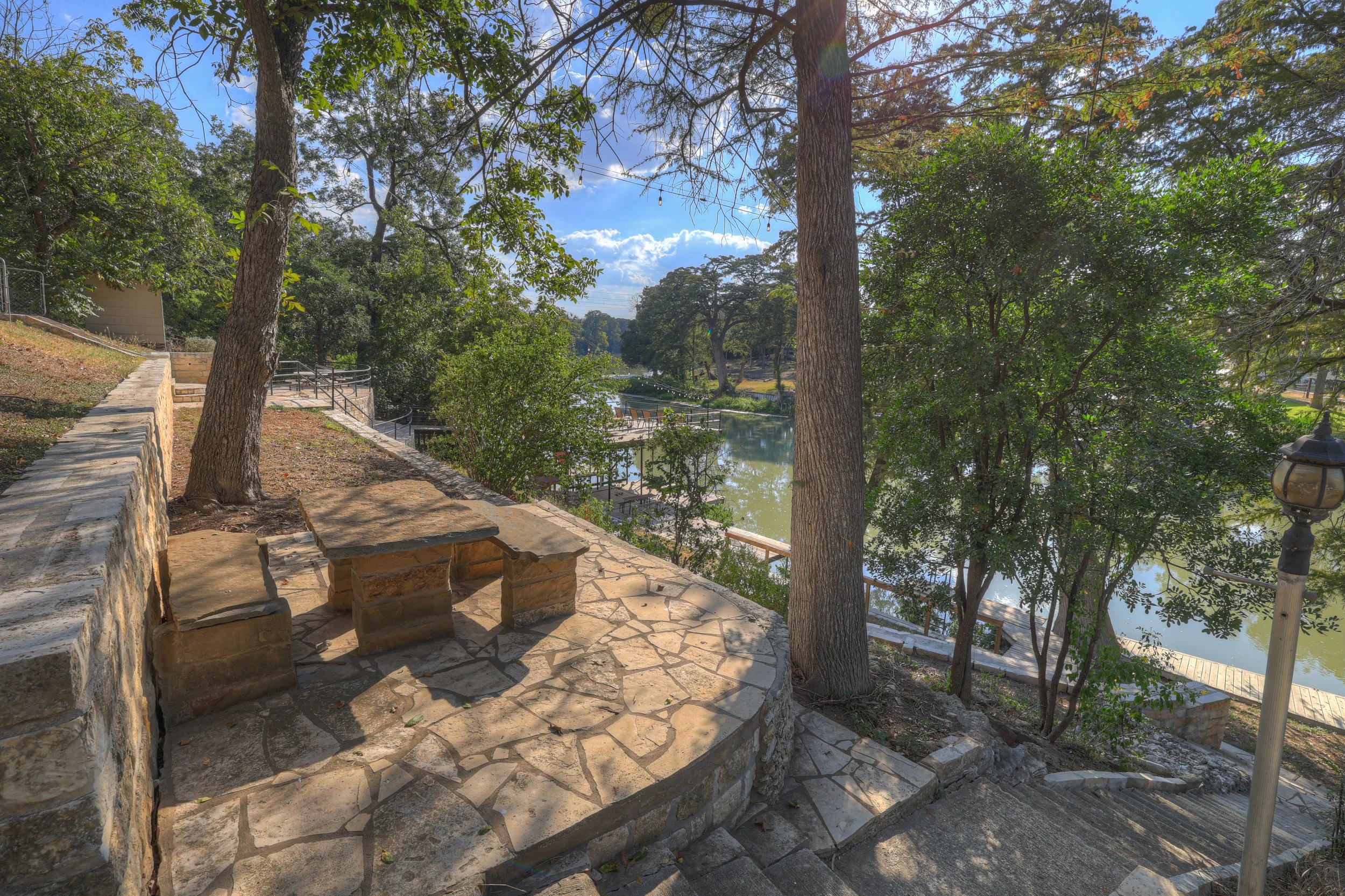 A stone pathway with a stone bench, trees providing shade, and a view of a river or lake through the trees. There is a lamppost on the right side, and the sky is partly cloudy with sunlight filtering through the trees.